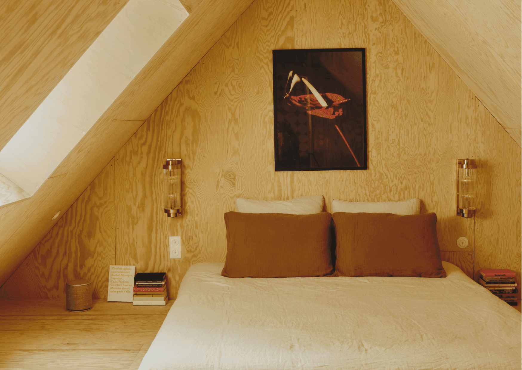 A cozy attic bedroom features slanted wooden walls and a skylight. The focal point is a bed with white bedding and brown pillows. On the wall behind the headboard, there’s an abstract painting from Maison Sauvage. Next to the bed are two small shelves holding books and a cylindrical speaker.