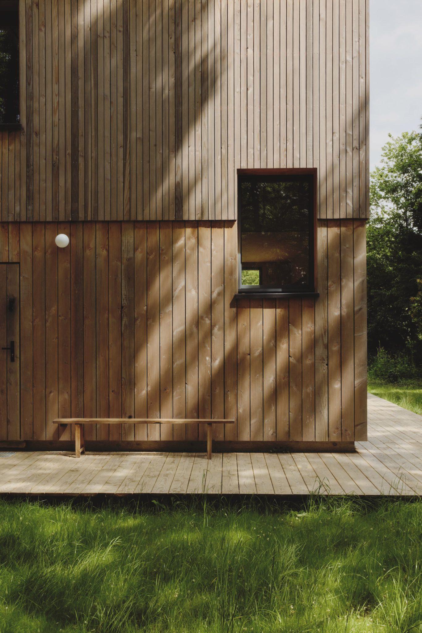 A modern wooden building with vertical slats and a small window is partially in shadow. A plank bench is positioned on the wooden terrace in front of the building. Green grass and trees are visible, suggesting a natural outdoor setting at Maison Sauvage.