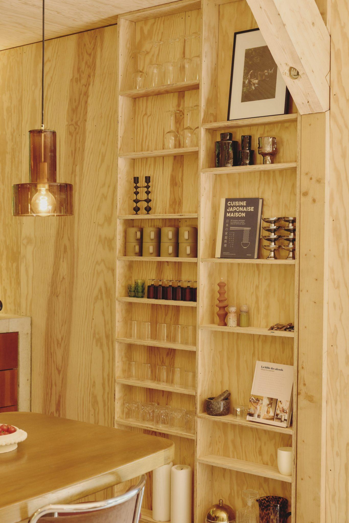 Un intérieur cosy en bois avec une bibliothèque intégrée à la Maison Sauvage. Les étagères sont remplies de verrerie, d'objets de décoration et de livres. Une photo encadrée en noir et blanc se trouve sur l’étagère du haut. Une suspension à la chaude lueur ambrée est suspendue au-dessus d’une table en bois au premier plan.
