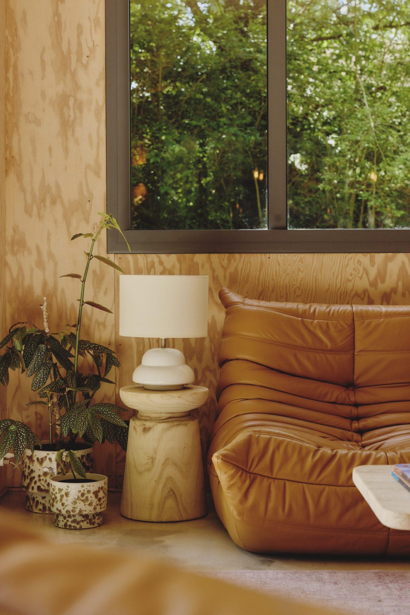 Un coin confortable dans une pièce moderne comprend un canapé capitonné en cuir beige, une table d'appoint en bois avec une lampe Maison Sauvage blanche et des plantes en pot. Une grande fenêtre offre une vue sur les arbres verts luxuriants à l'extérieur, ajoutant à l'atmosphère sereine.