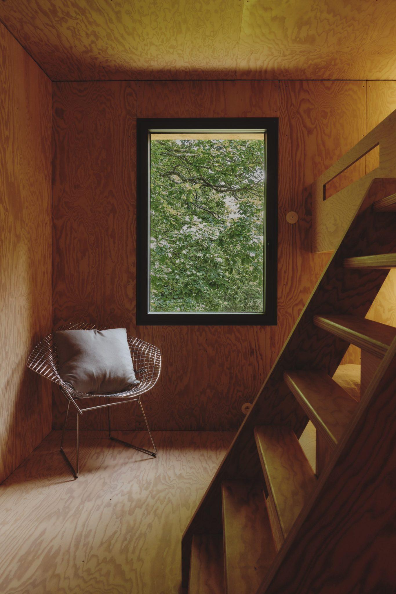 A cozy wooden corner features a wire chair with a cushion next to a large window overlooking green trees. A small staircase with wooden steps leads up to a higher level, capturing the peaceful and natural ambiance of Maison Sauvage.