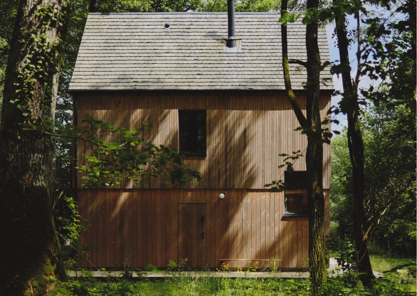 Une cabane en bois de deux étages avec un toit en bardeaux en pente, nommée Maison Sauvage, est située au milieu de grands arbres et d'une verdure luxuriante. Le bâtiment présente un design minimaliste avec quelques fenêtres et une porte. La lumière du soleil filtre à travers les arbres, projetant des ombres sur l'extérieur en bois de la cabane.