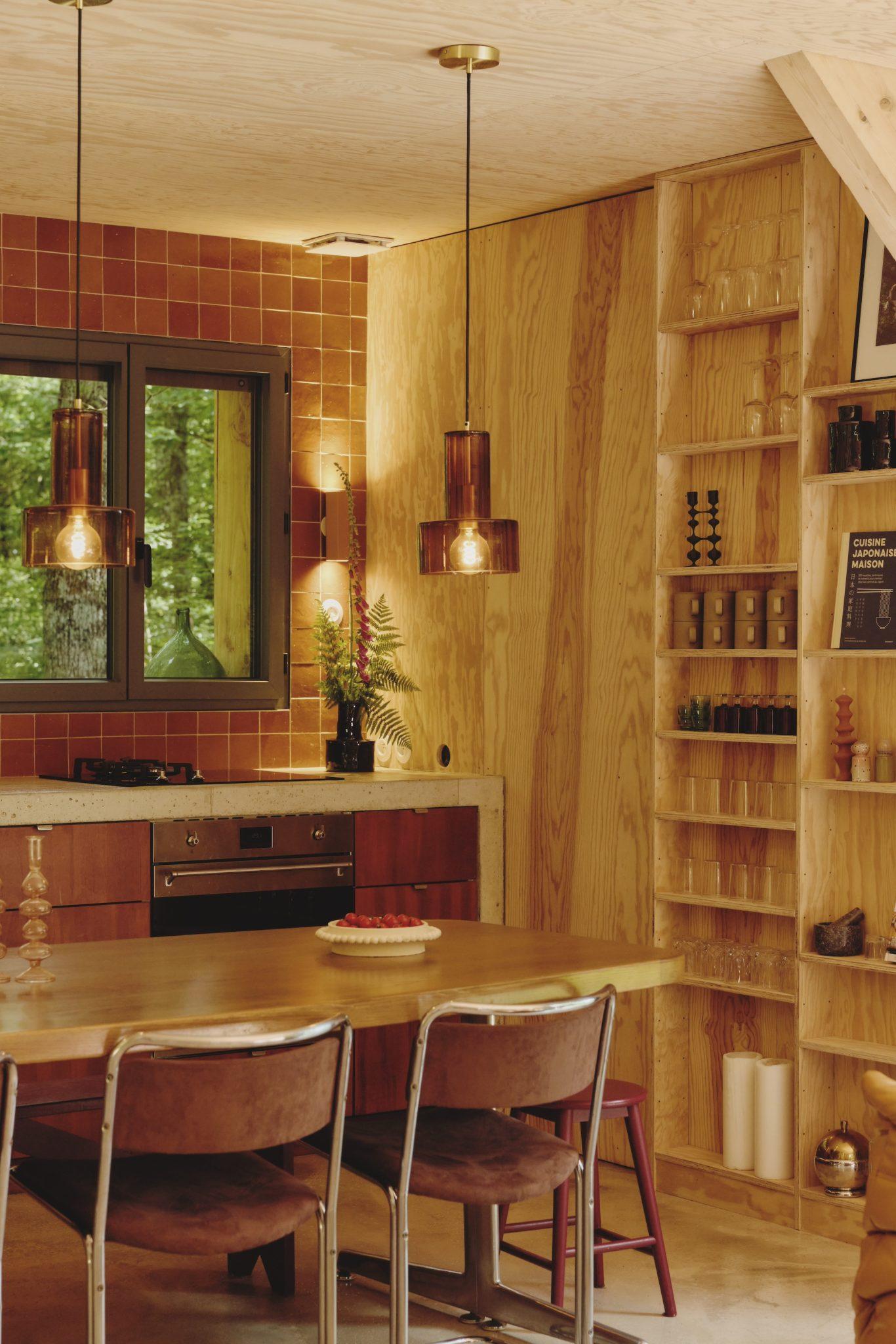 A cozy kitchen and dining area from Maison Sauvage features a wooden table with four chairs, pendant lights, a tiled backsplash, and open shelves. The room is adorned with indoor plants, and a window provides a view of the greenery outside. A cake is placed on the table.