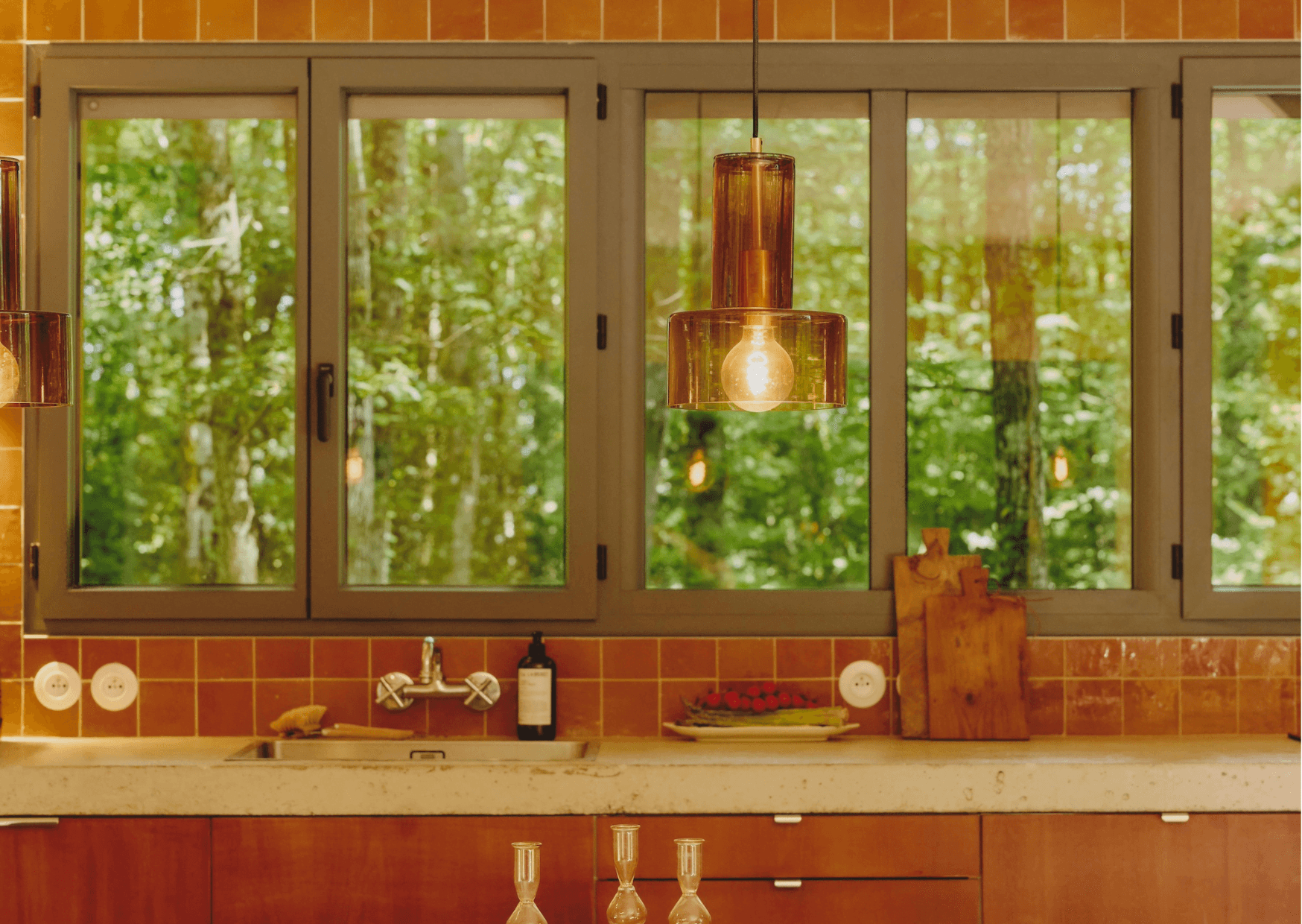 A cozy kitchen from Maison Sauvage featuring wooden cabinets, a light countertop, and a backsplash of earth-toned tiles. Large windows offer a view of a lush forest. A pendant light hangs above, and various kitchen items, including wooden cutting boards, are neatly arranged on the countertop.