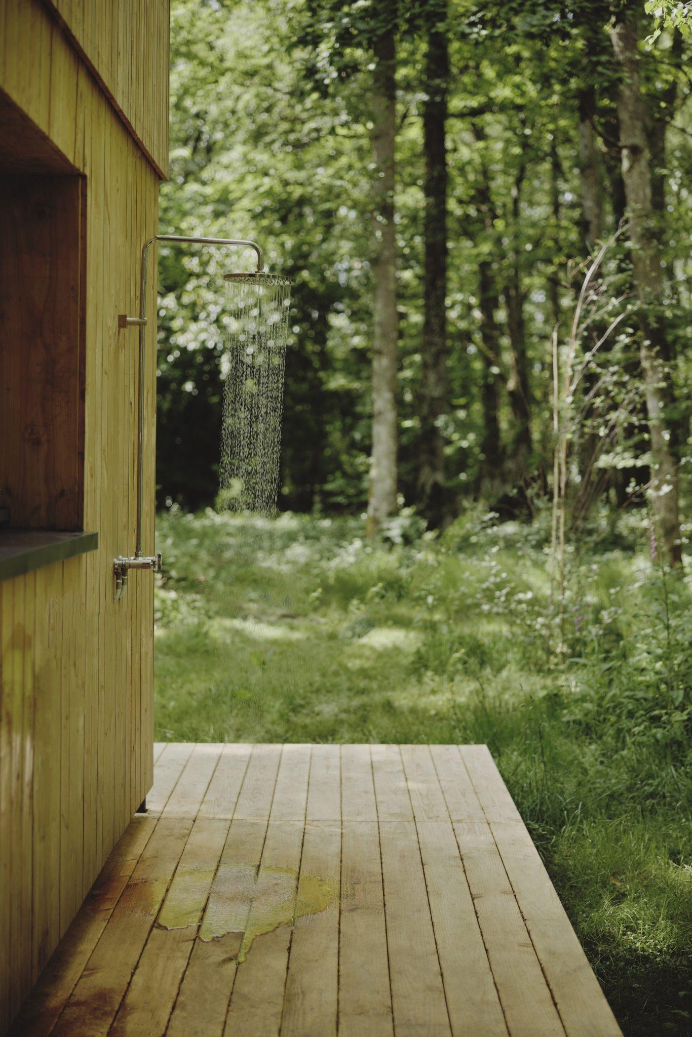 An outdoor shower, designed by Maison Sauvage, is installed on the side of a wooden building in a lush forest setting. Wooden planks create a small deck in front of the shower, while trees and greenery are visible in the background, creating a serene and natural atmosphere.