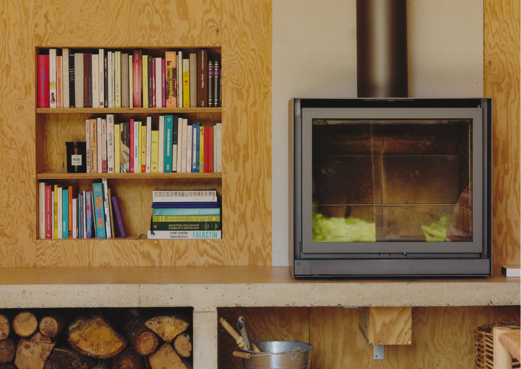 Un espace de vie comprenant une bibliothèque en bois remplie de livres colorés à gauche, une cheminée moderne avec une façade en verre à droite et du bois de chauffage empilé en dessous. Un seau en métal avec des outils de cheminée se trouve devant le bois de chauffage, ajoutant au charme rustique de cette pièce d'inspiration Maison Sauvage.