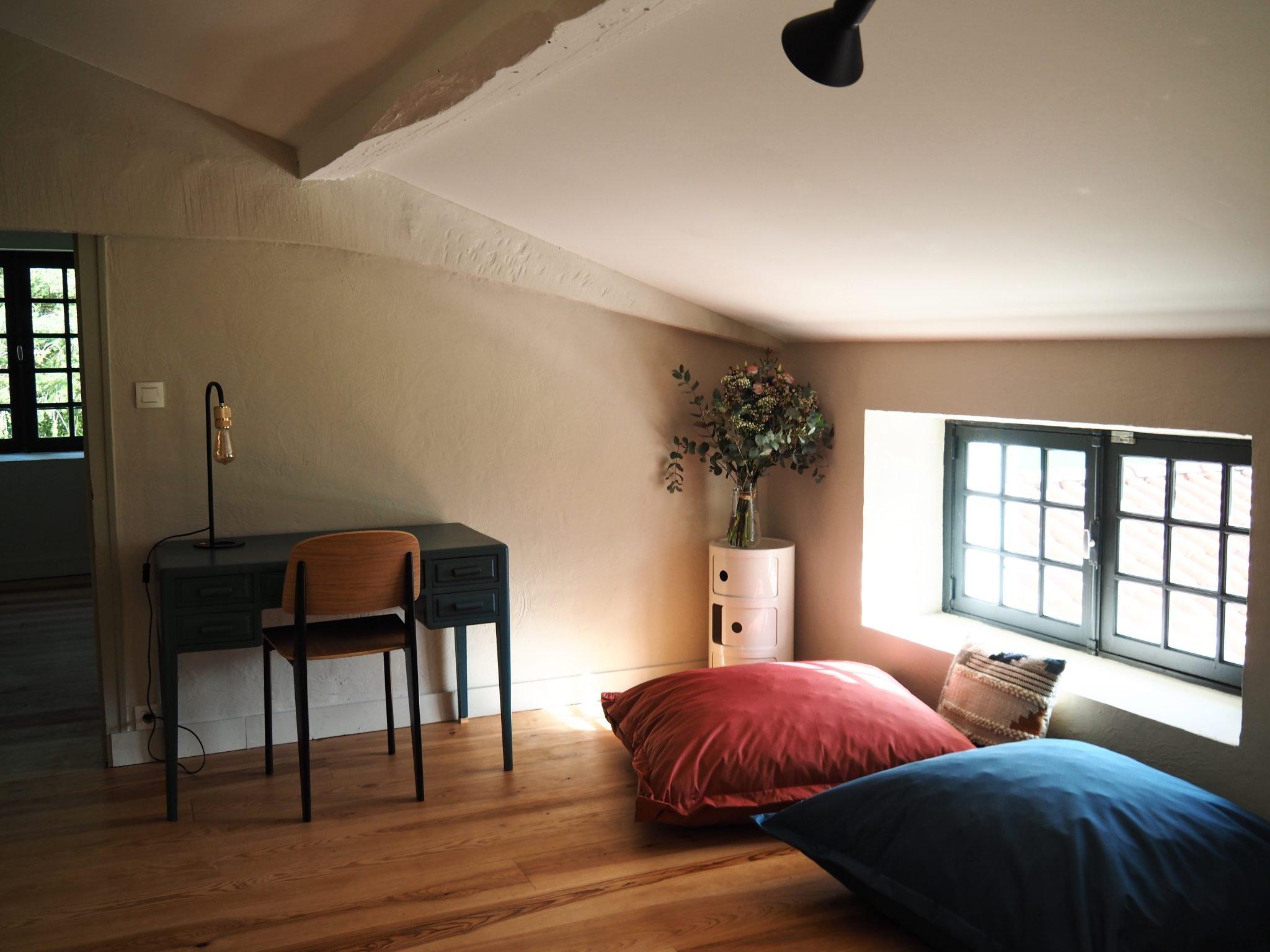 A cozy attic bedroom with sloped ceilings features a small desk and chair against the wall, a floor lamp, a round white wardrobe with a vase of flowers, and two large floor cushions in blue and red near a small window overlooking the rooftops.