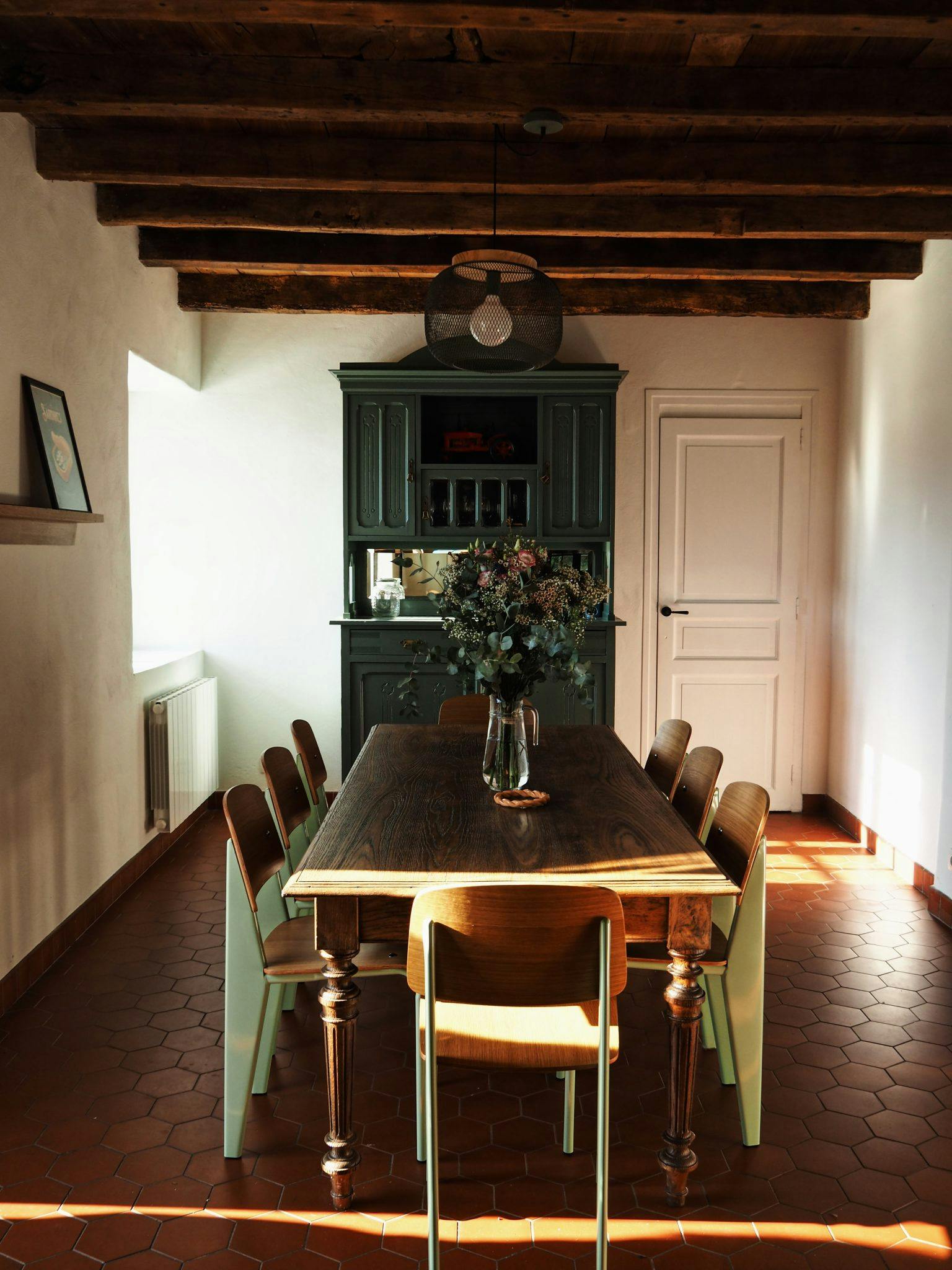 A cozy dining room with wooden beams, a rustic wooden table, light green chairs, and a vase of flowers on the table. The room features a green cabinet against the back wall and a white door beside it. Sunlight filters in, creating a warm and inviting atmosphere.