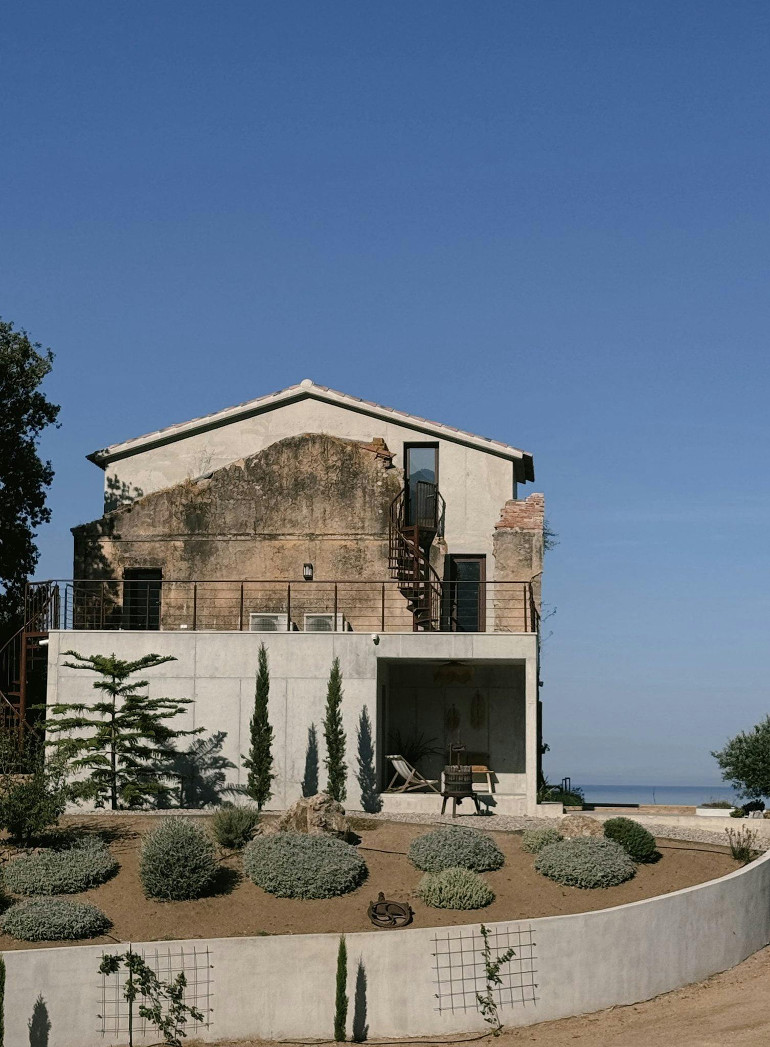 Une ancienne maison à deux étages avec une façade patinée par le temps et un niveau inférieur moderne, comprenant un balcon, un coin salon extérieur et un jardin décoratif avec des plantes et des arbustes variés. Cette destination de luxe en Corse offre le décor parfait d'un ciel bleu clair et d'un aperçu lointain de la mer.