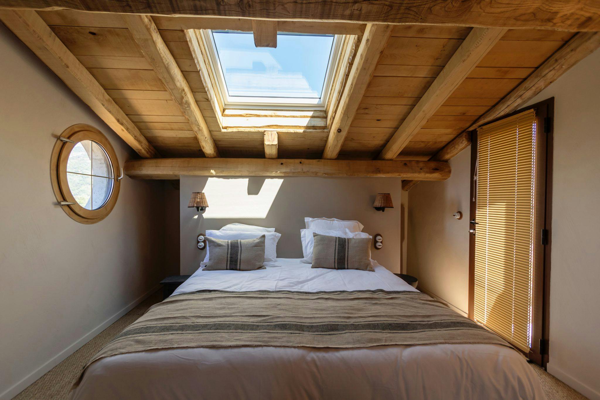 A cozy attic bedroom with wooden beams on the ceiling, a skylight, and a round window. The room features a neatly made double bed with white linens and striped pillows. On the right, there are wooden blinds covering a window, casting a warm light across the space.