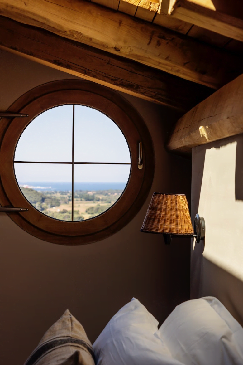 Round window in the bedroom overlooking the mountains. Bed, nightstand, and small bedside lamp.