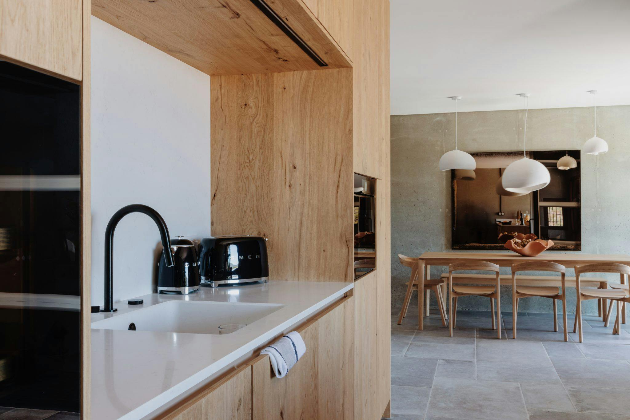 A modern kitchen and dining area with wooden cabinetry and light fixtures. The kitchen features a white countertop with a black faucet, a black toaster, and a coffee maker. The dining area has a large wooden table with light wooden chairs and pendant lights above.