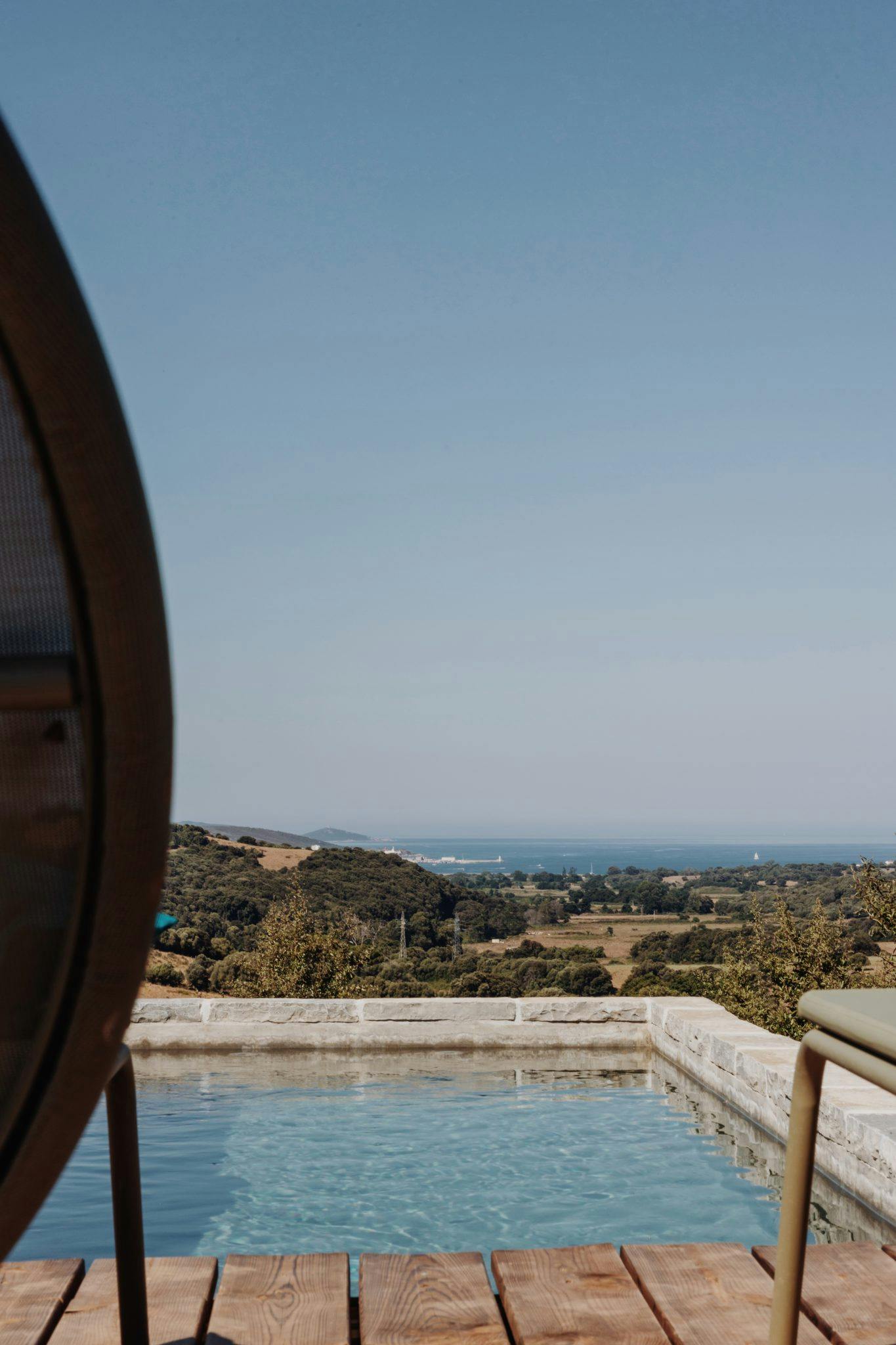 Une vue pittoresque d'un paysage serein avec un ciel bleu clair, une vallée verdoyante et un horizon bleu lointain. Au premier plan, une terrasse en bois entoure une piscine, partiellement visible derrière un mobilier d'extérieur. La scène respire la tranquillité et la détente.