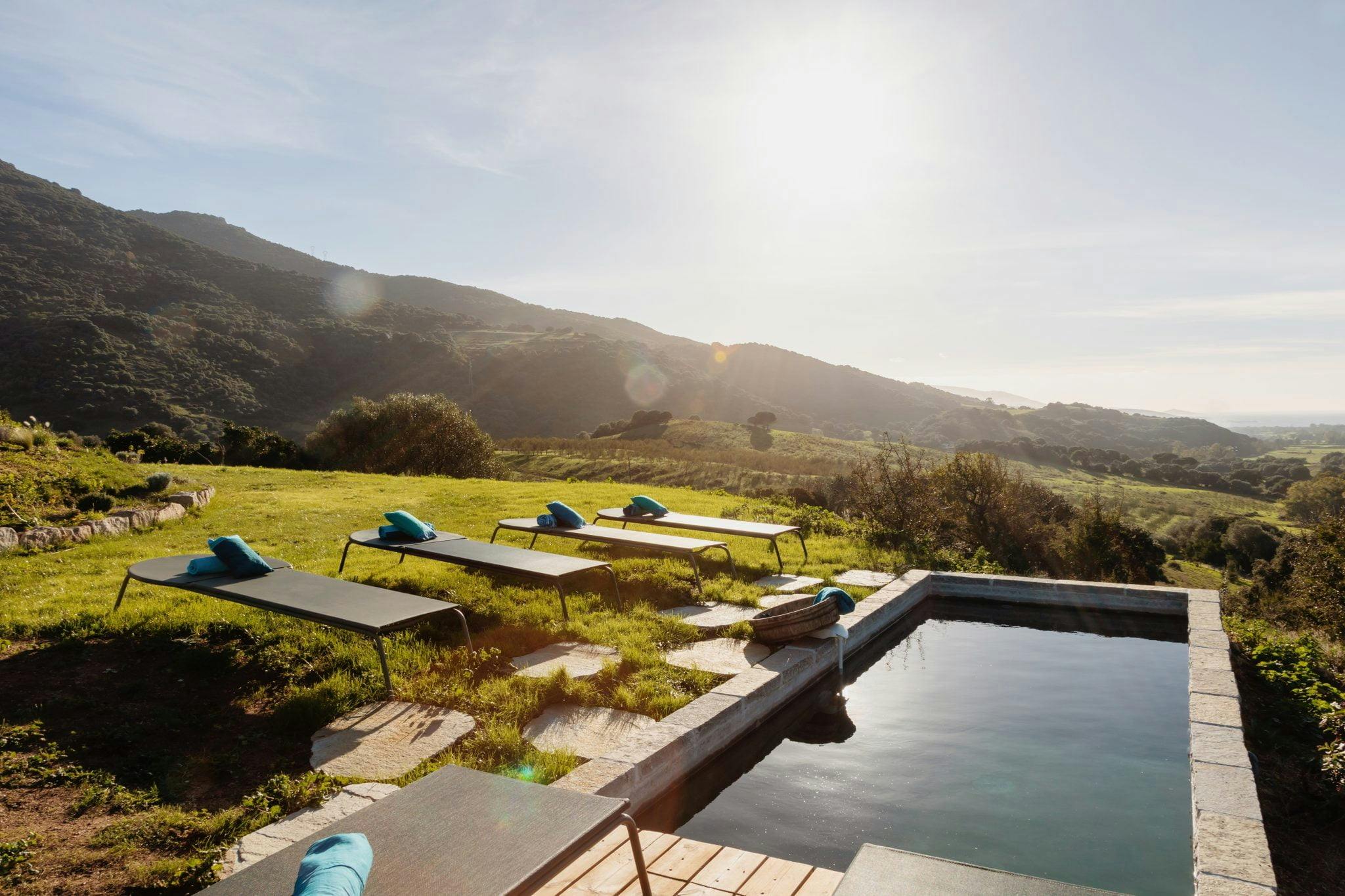 A serene outdoor setting featuring a pool with stone edges overlooking a lush green valley and distant mountains under a bright, clear sky. Four lounge chairs with blue cushions are placed around the pool, inviting relaxation and enjoyment of the natural scenery.