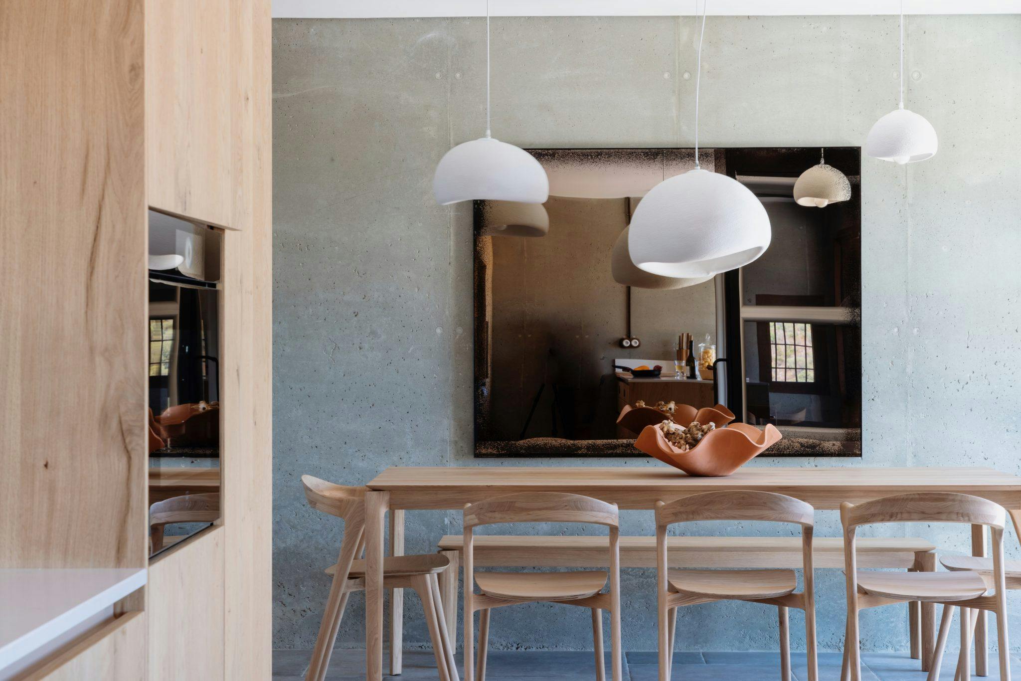 Une salle à manger moderne comprend une table en bois avec six chaises assorties et un centre de table unique en céramique. Au-dessus, trois suspensions blanches sont suspendues. Un grand miroir réfléchissant recouvre le mur du fond, ajoutant de la profondeur à la pièce au design minimaliste.