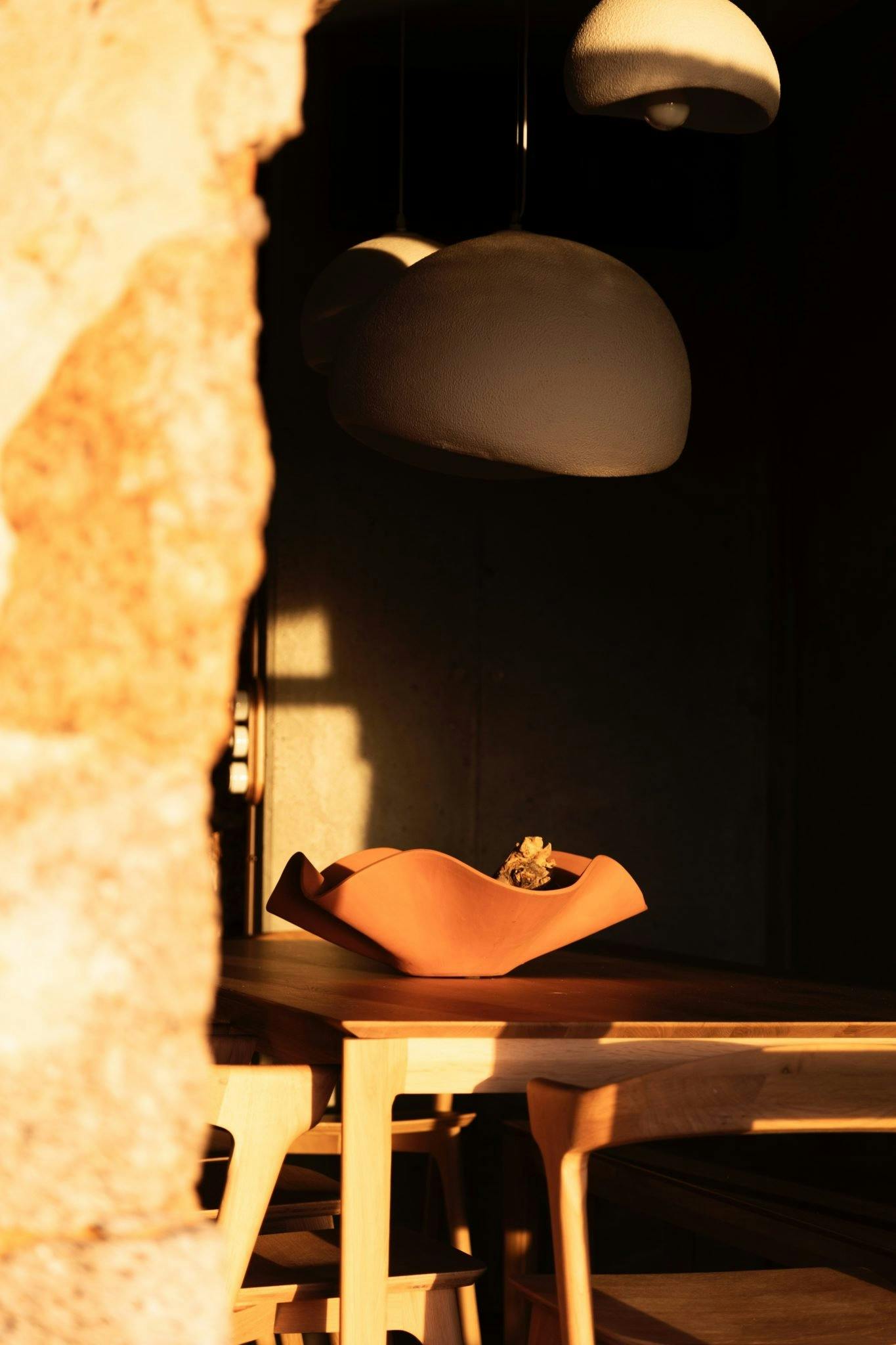 A warm sunlit room with modern decor featuring a wooden table and beige chairs. The table has a unique, wavy terracotta bowl with a small decorative item inside. Overhead, there are three large, dome-shaped light fixtures in a minimalist style.