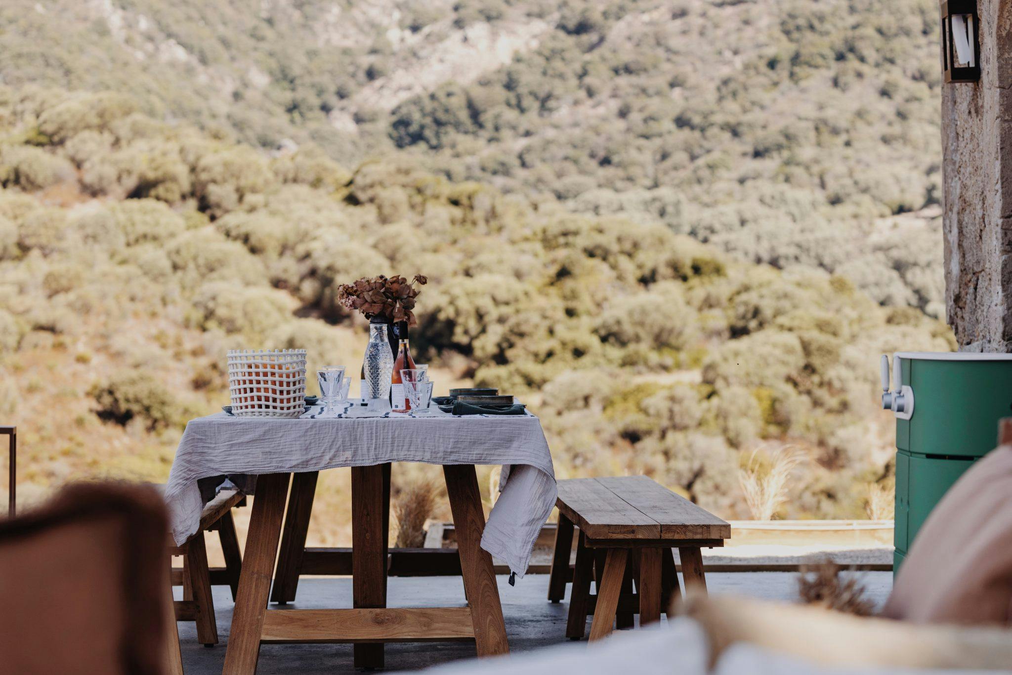 Une salle à manger rustique en plein air comprend une table et des bancs en bois avec une nappe légère, des fleurs séchées et de la vaisselle. La toile de fond est une vue panoramique de collines verdoyantes et luxuriantes sous un ciel clair. Une atmosphère chaleureuse et accueillante est créée pour les repas en plein air.