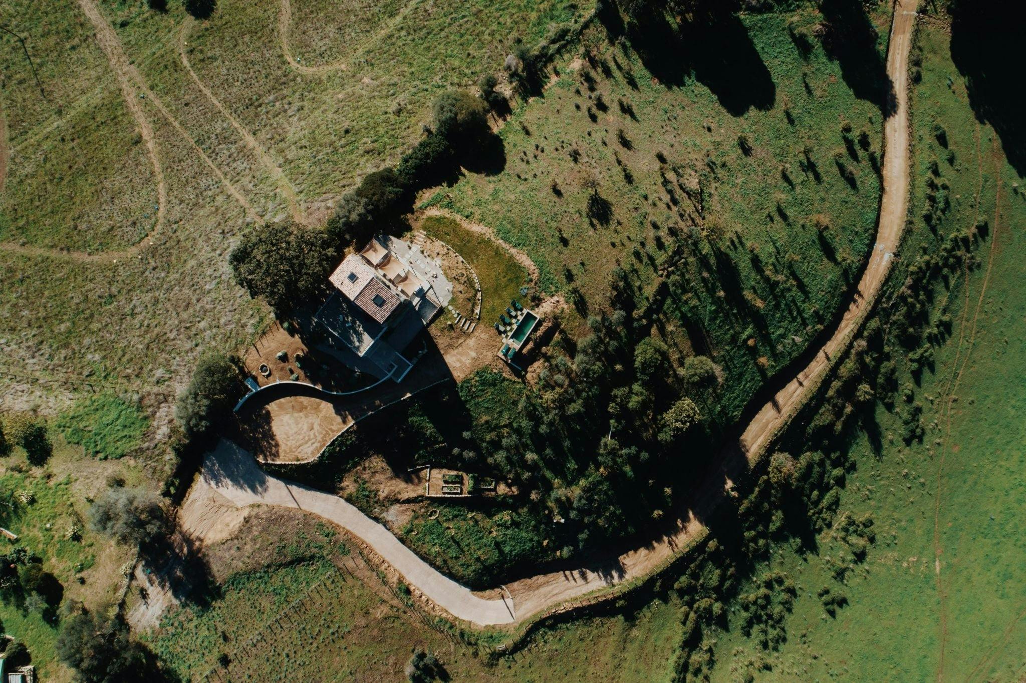 Vue aérienne d'un paysage rural présentant une maison solitaire entourée de verdure. Un chemin de terre sinueux serpente à côté de la maison, traversant des champs et des arbres épars. La zone présente un mélange d’espaces herbeux ouverts et de grappes de végétation.