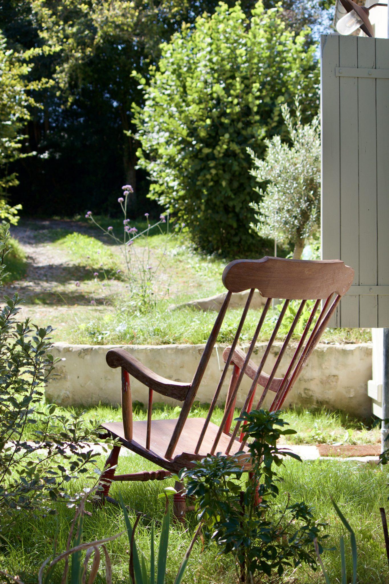 Une chaise à bascule en bois est placée sur une pelouse dans un jardin ensoleillé. La scène est entourée de diverses plantes vertes, et il y a un mur blanc et une porte blanche ouverte à droite. L'atmosphère générale est paisible et accueillante.