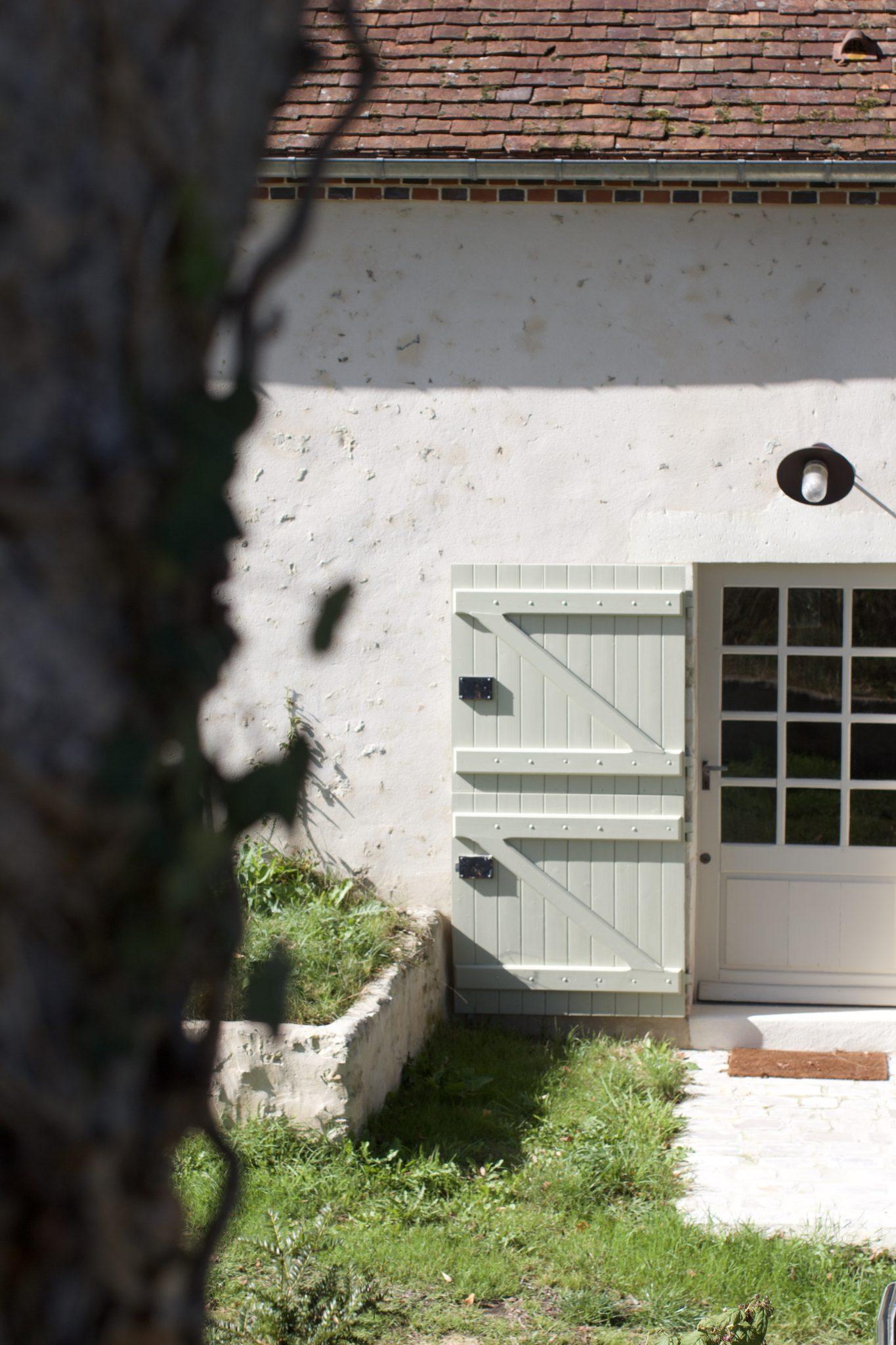 Un bâtiment rustique en stuc blanc avec un toit de tuiles en pente et une petite fenêtre dans la partie supérieure. Une porte ouverte en bois vert pâle est fixée à la moitié inférieure d’une porte vitrée. Au premier plan, un arbre obscurcit partiellement la vue.