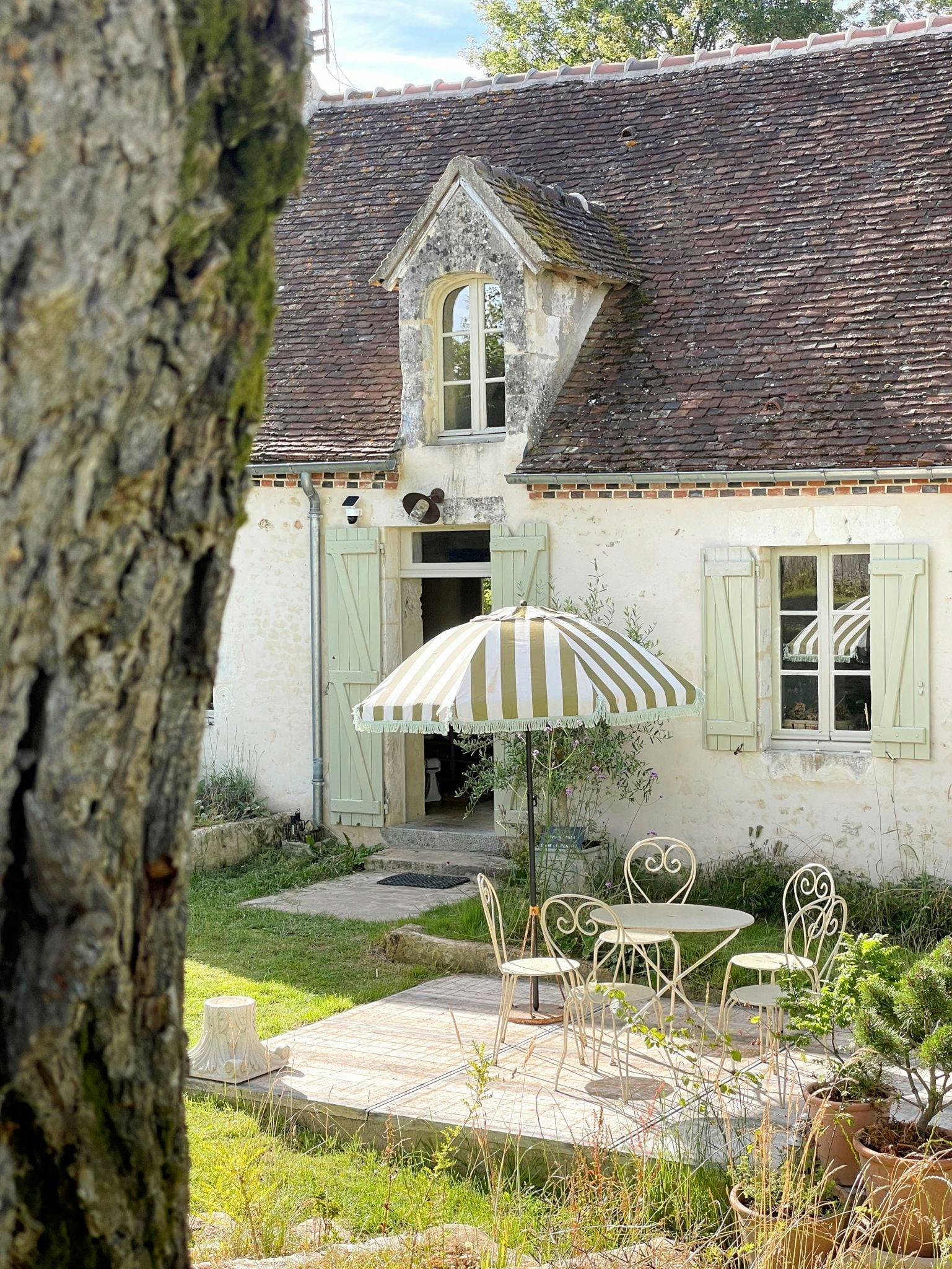 A charming rustic cottage with a steeply pitched roof and light-colored shutters. In front of the house is a small patio with a round table, matching chairs, and a green and white striped umbrella. A tree trunk frames the left side of the image.