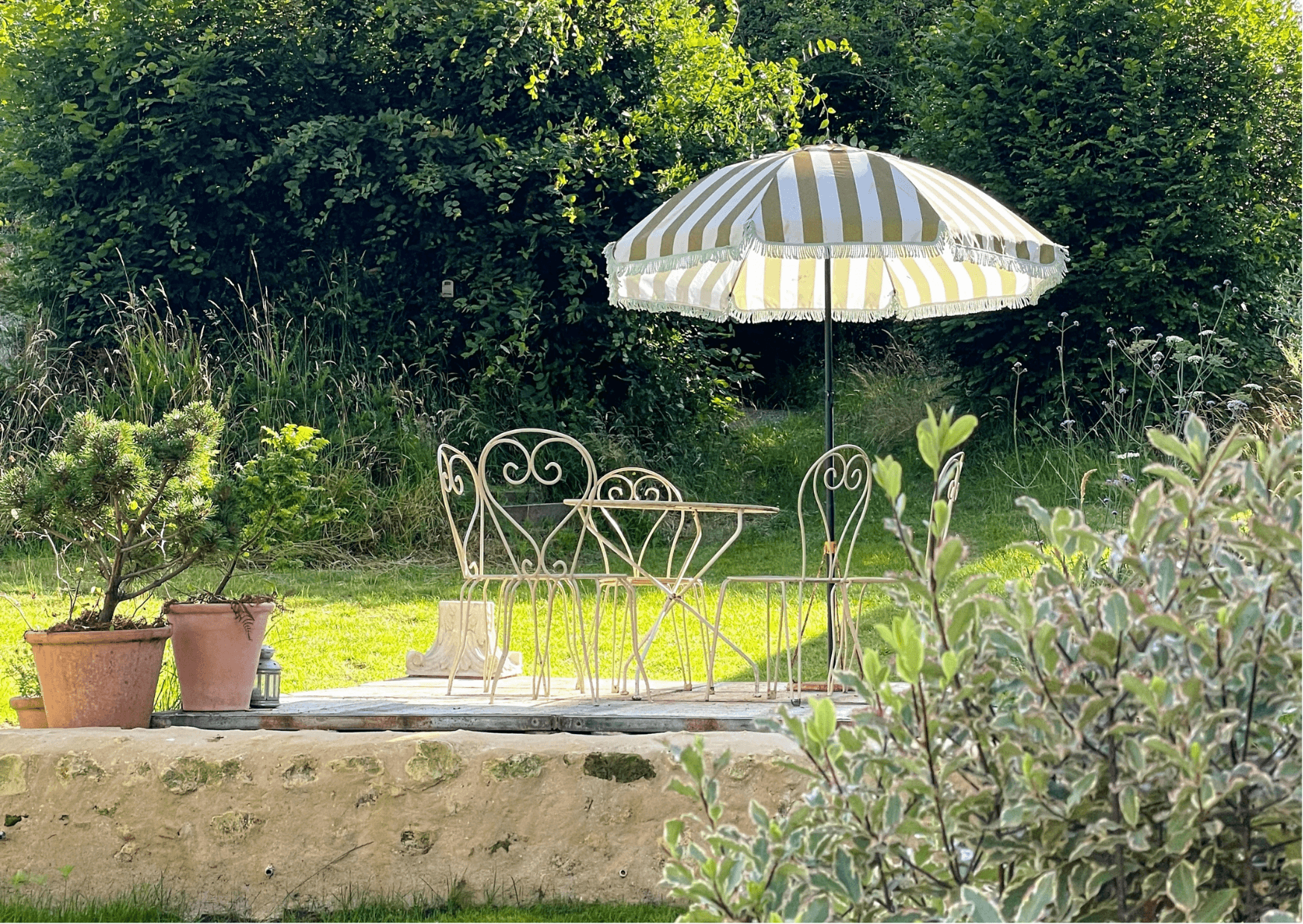 A picturesque outdoor seating area featuring a wrought-iron table with matching chairs under a white and yellow striped umbrella, surrounded by lush greenery and potted plants on a sunny day.