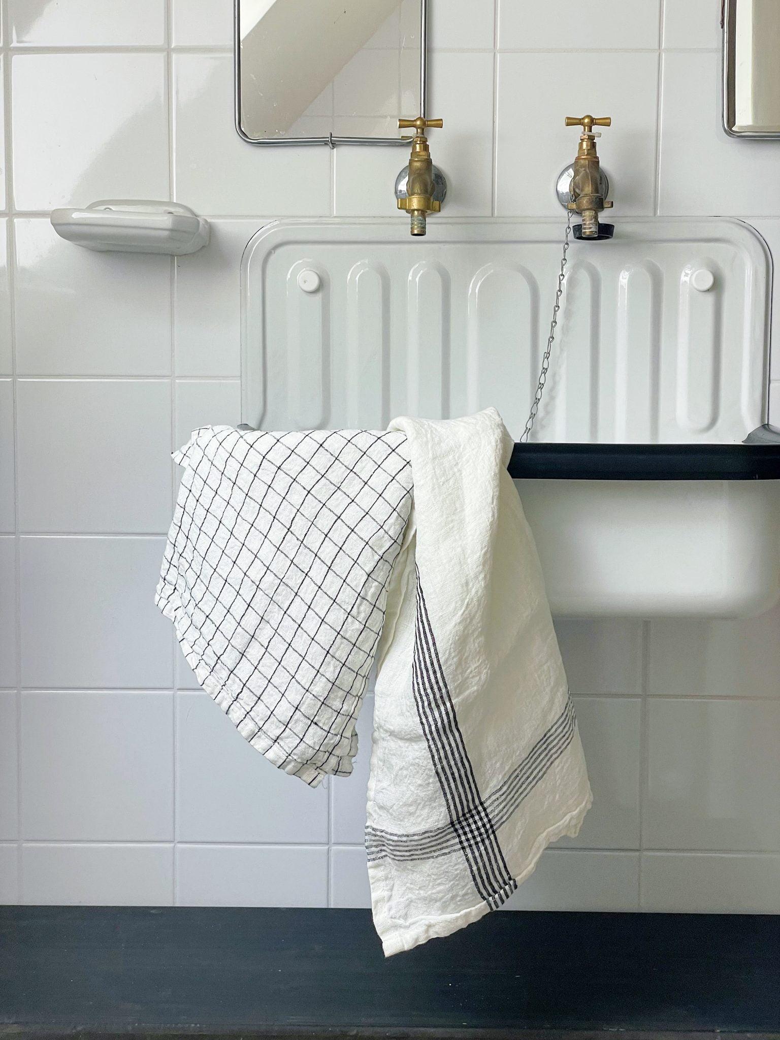 A white tiled wall with a rectangular white sink featuring two brass faucets. A white towel with a black checkered pattern is draped over the edge of the sink. The sink has a built-in soap dish in the wall and a chain attached to the sink plug. The space appears clean and minimalist.