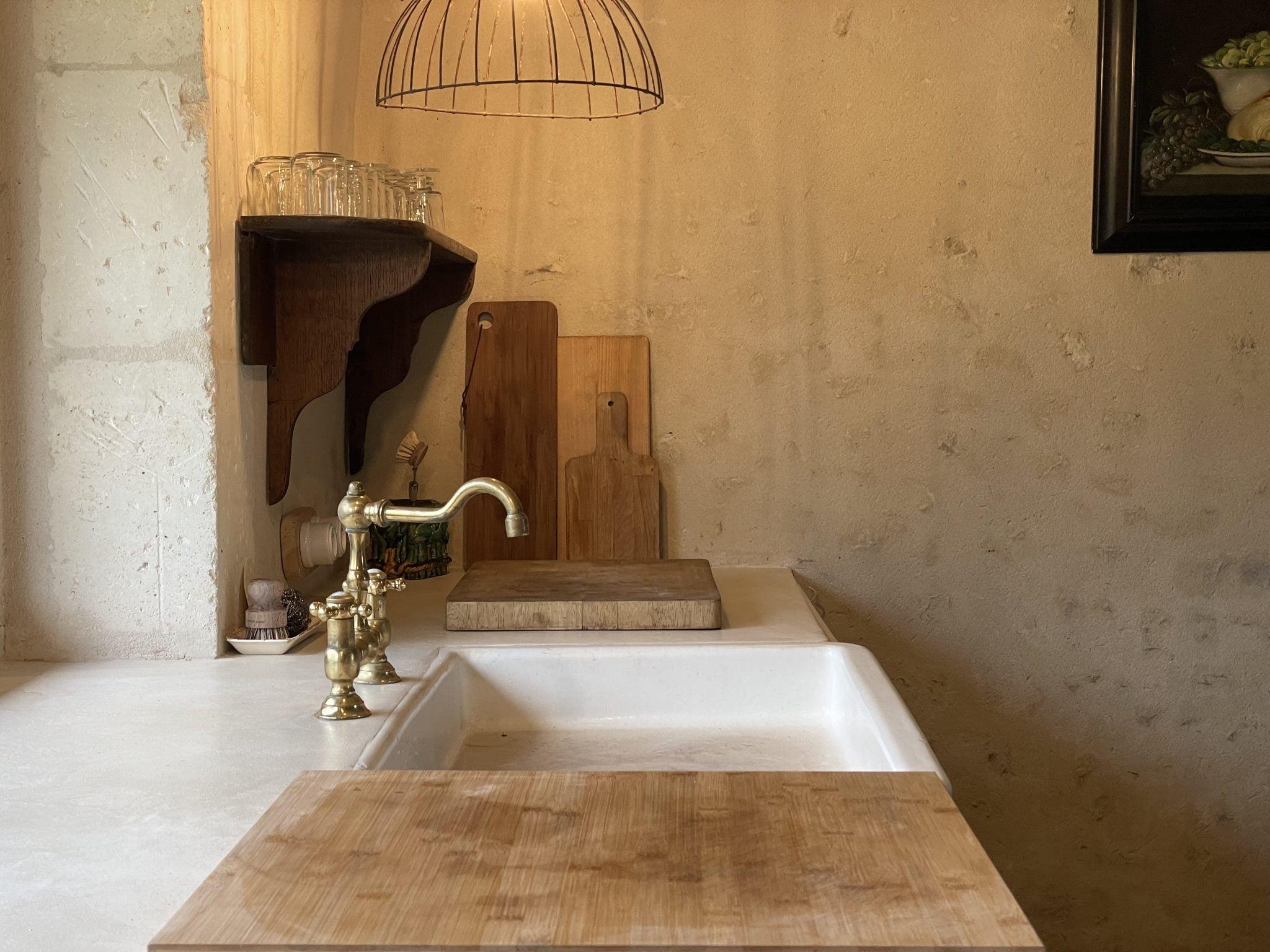 A rustic kitchen counter features a brass faucet over a white sink, with wooden cutting boards and glasses on a wooden shelf. A wire pendant light hangs above the counter, and the wall has a textured, raw, and unfinished look.