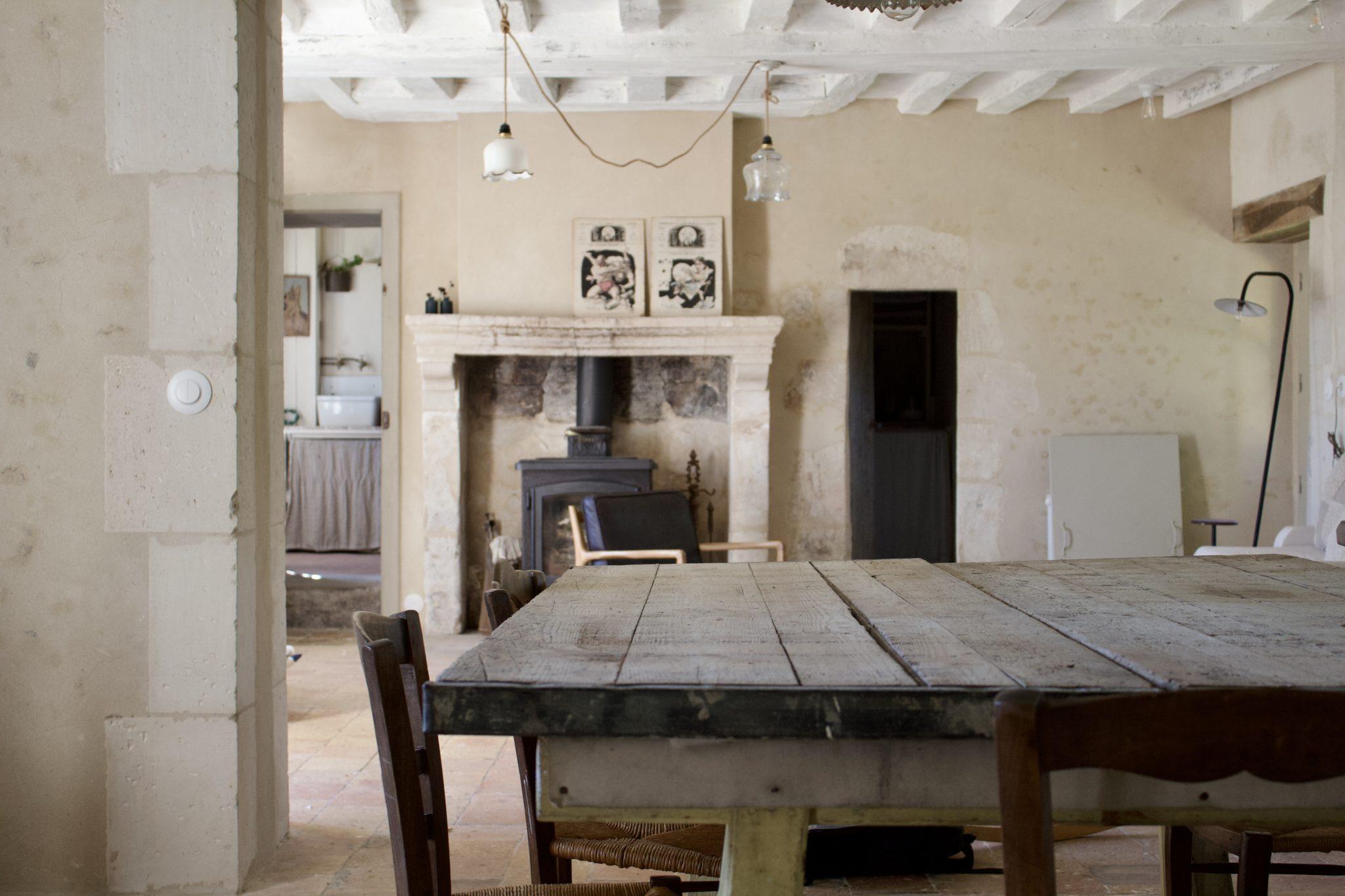 A rustic room with a wooden dining table and several chairs. In the background, there is a fireplace with a wood-burning stove, framed artwork, and exposed beams on the ceiling. A door leads to another room with shelves and visible decorative items.