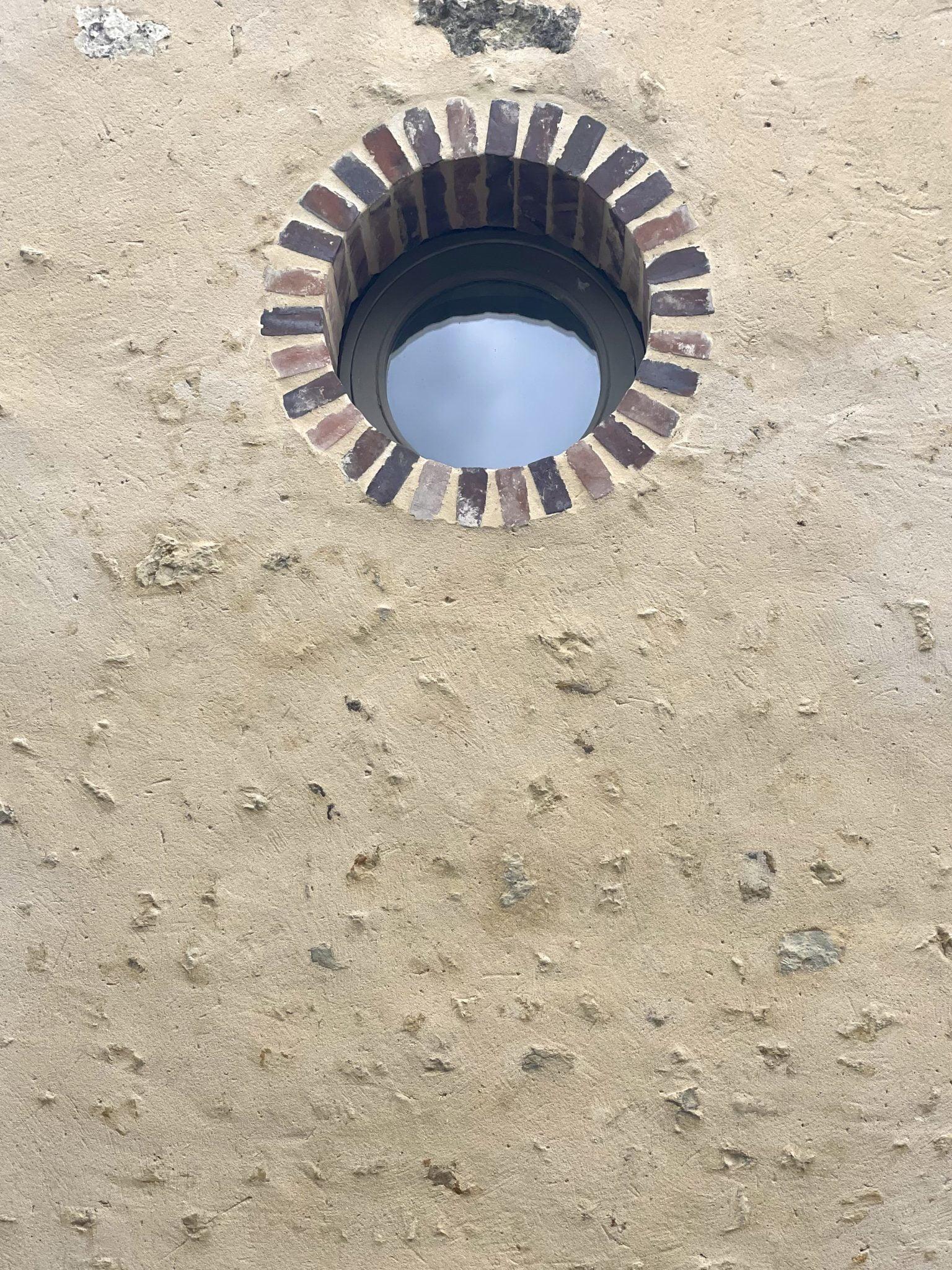 A round window with a brick surround is set into a light-colored textured wall. The wall features various small, unevenly spaced indentations, giving it a rustic appearance. The glass of the window reflects the sky.