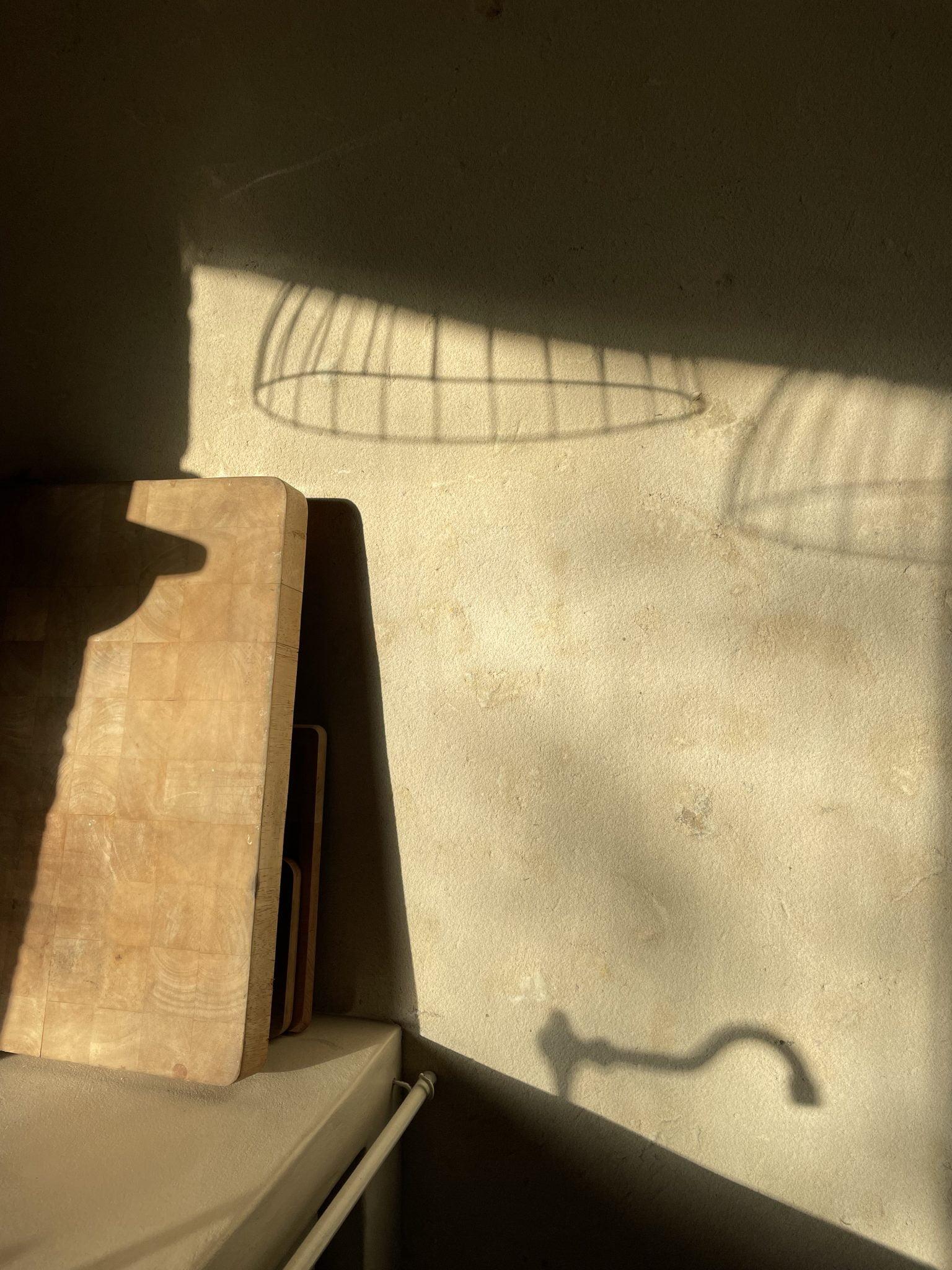 The shadow of a metal wire lampshade and a hanging faucet is cast on a textured wall bathed in sunlight. Beneath the shadow, wooden cutting boards are propped against the wall next to a countertop. The overall scene has a warm and rustic ambiance.