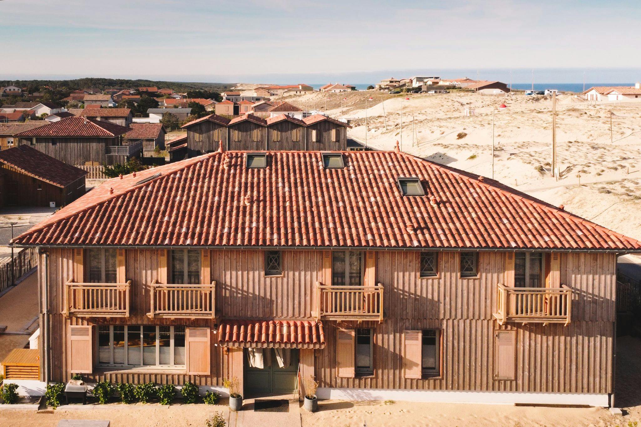 Facade of a large wooden house on the dunes, with balconies and the sea in the background.