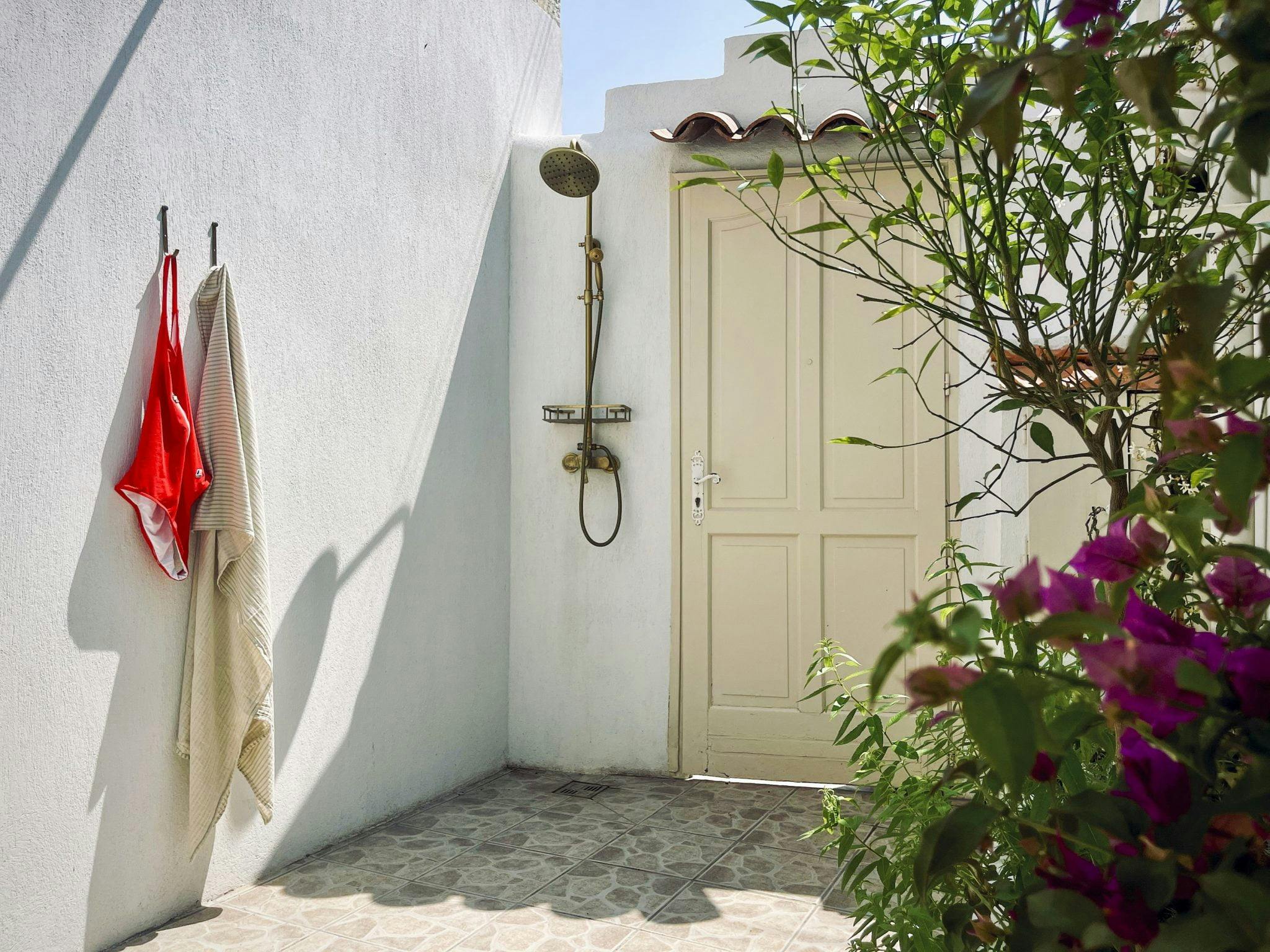 An outdoor shower area with a beige door and a mounted showerhead against a white wall. A red and white towel hangs on hooks on the left. Purple flowers and green foliage on the right add a touch of nature. The ground is tiled, and sunlight illuminates the space.