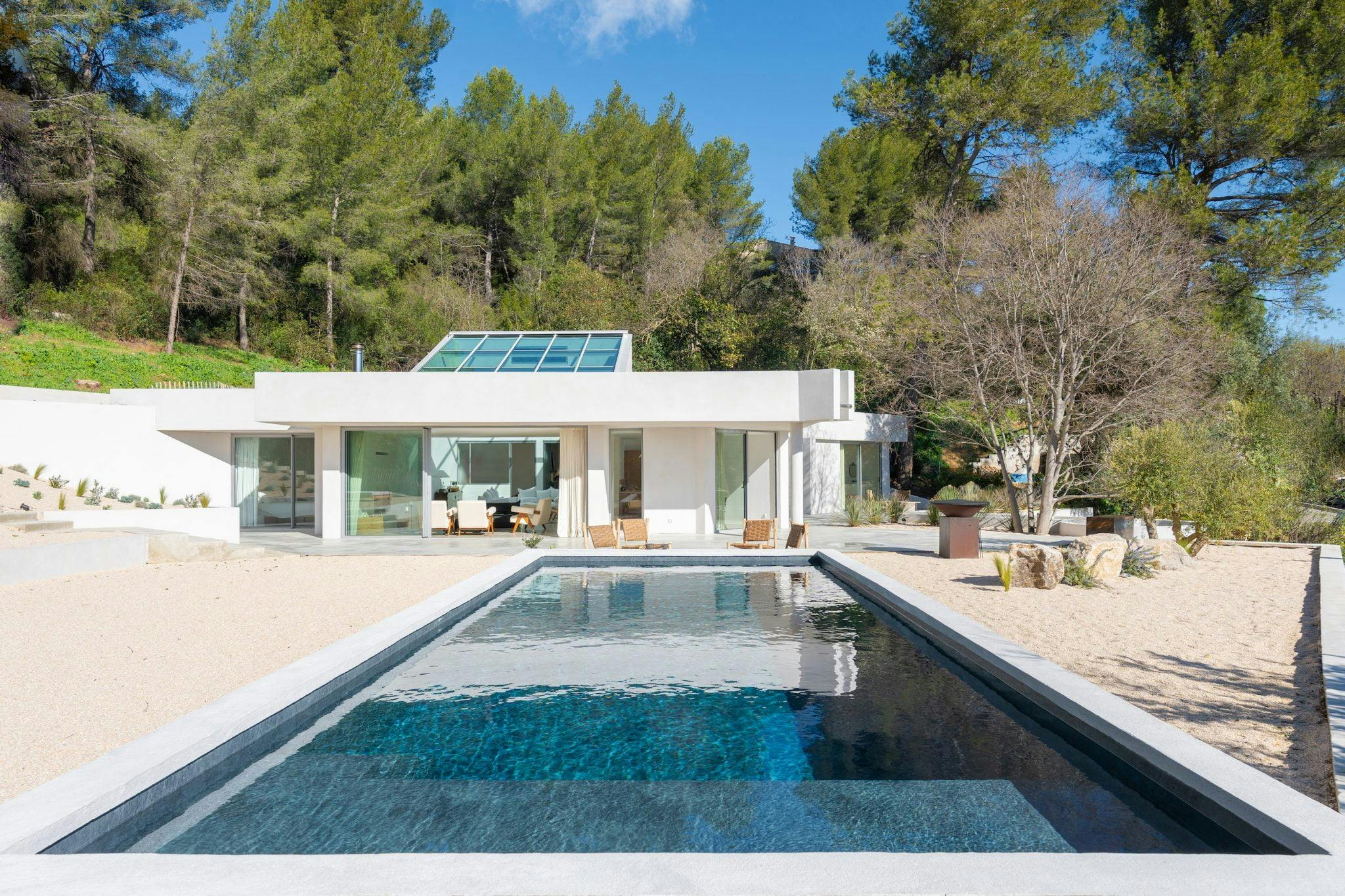 Modern white house with large windows and glass doors, surrounded by trees. It features a long rectangular pool extending towards the house, lounge chairs by the pool, and a minimalist landscape with gravel paths. Clear blue sky above.