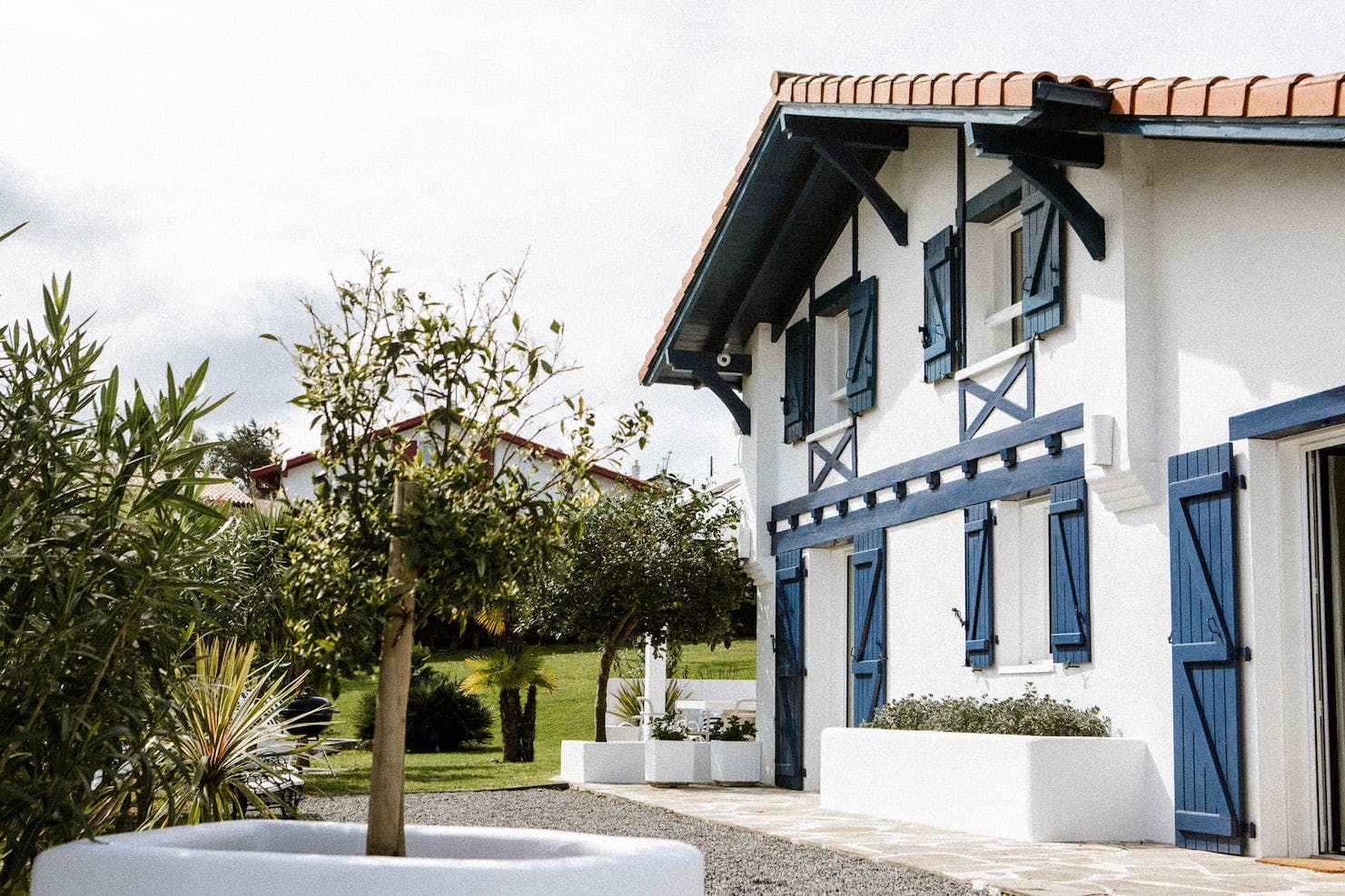 Une maison blanche à deux étages avec des volets et des boiseries bleues, avec un toit de tuiles rouges. La maison est entourée de verdure, dont un jeune arbre au premier plan et divers arbustes et plantes. Le ciel est partiellement nuageux. La zone est pavée de carreaux de pierre.