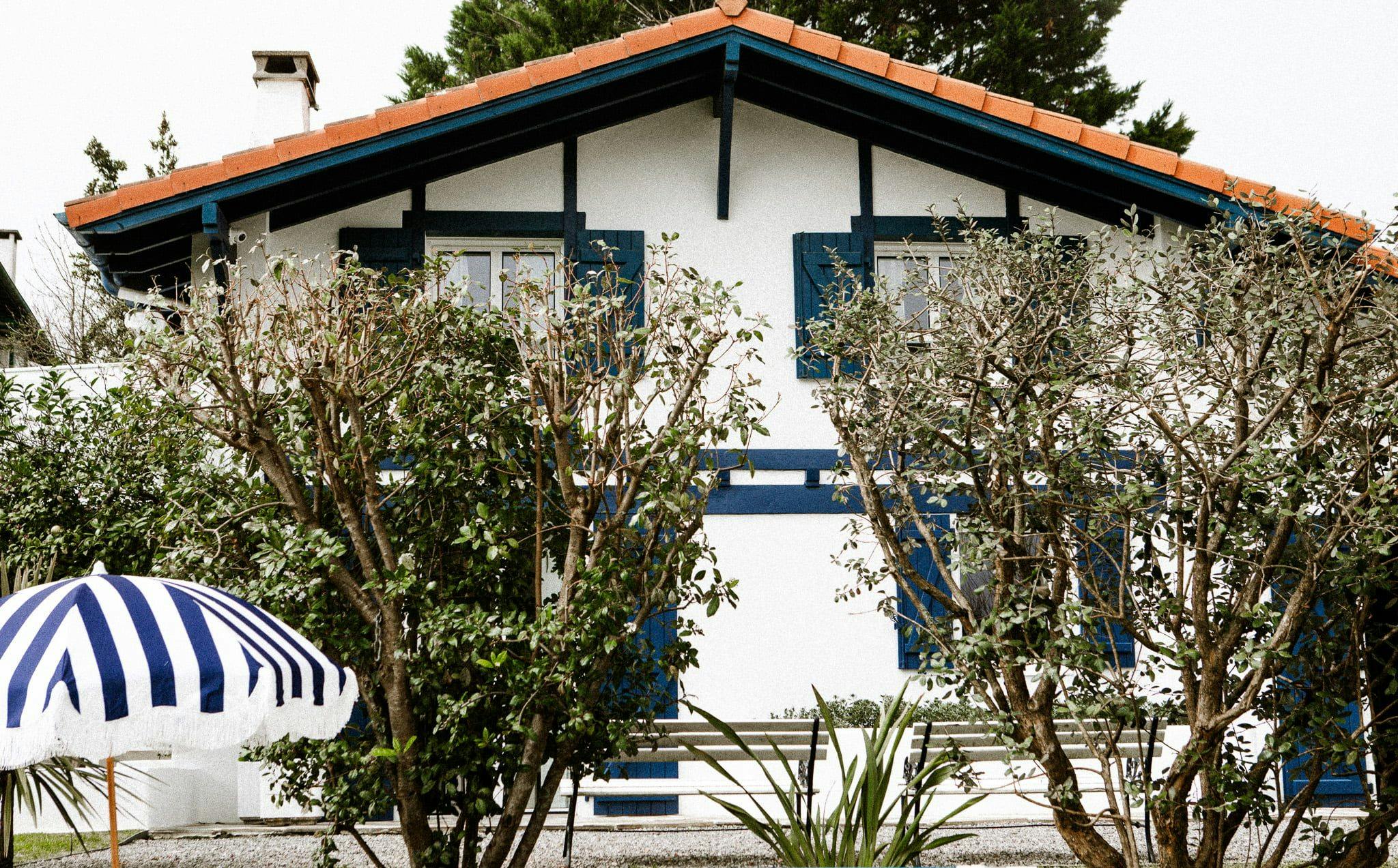 A charming two-story house with white walls, blue shutters, and a red-tiled roof is partially obscured by leafy trees and shrubs. A striped blue and white umbrella rests near a wall in the front yard of this Domino Un home, adding a cozy, inviting touch to the scene.