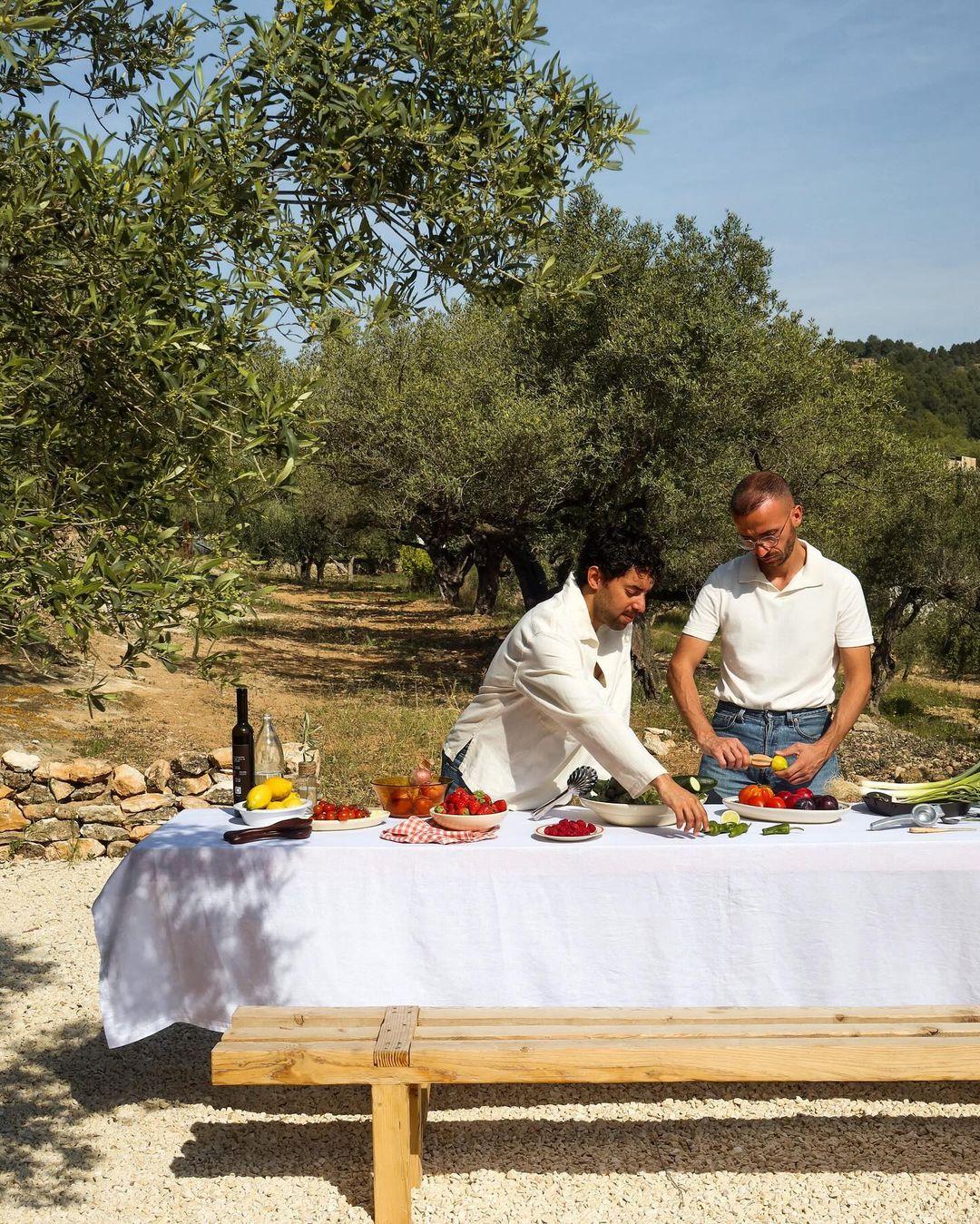 Deux hommes préparent un repas sur une longue table recouverte d'une nappe blanche, installée dans un décor extérieur entouré d'arbres et de verdure. Différents ingrédients et plats sont posés sur la table, ainsi qu'une bouteille de vin et des verres. Il semble que ce soit une journée ensoleillée.