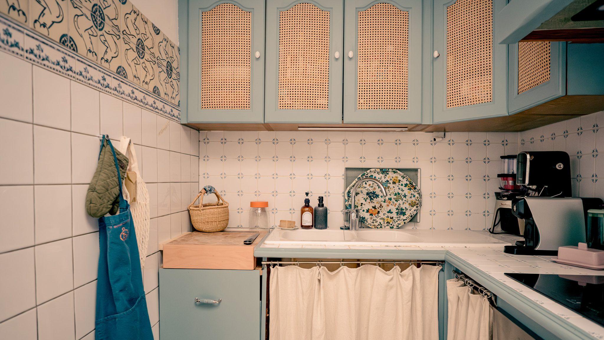 A small vintage-style kitchen with light blue cabinets featuring rattan panels. The countertop includes a woven basket, glass jars, and soap dispensers. A floral tray rests behind the sink, which has a curtain in front of it instead of cabinet doors.