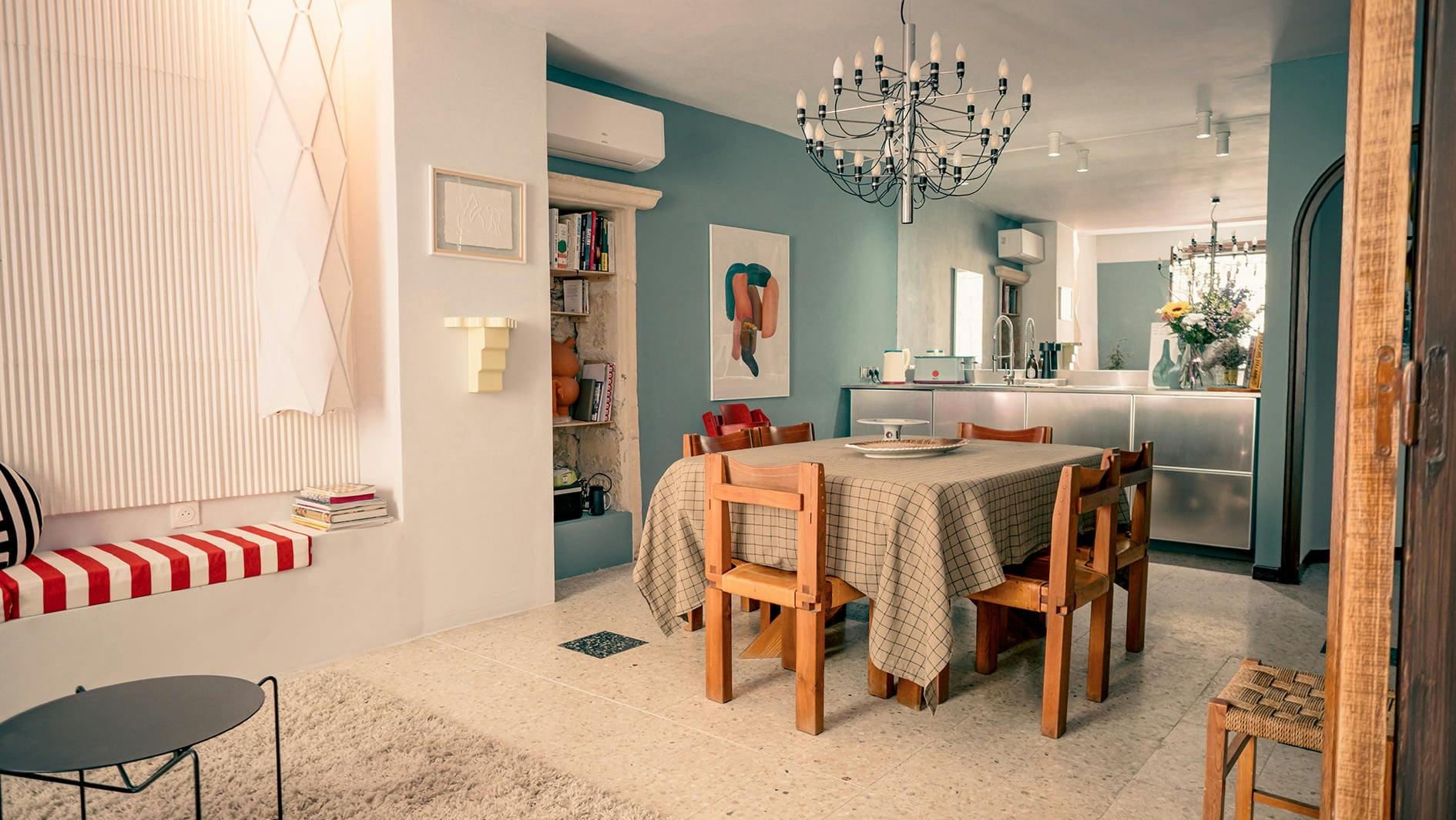 A cozy dining area with a wooden table and six matching chairs covered with a checkered tablecloth. The room features teal walls, a chandelier, a built-in bookshelf, and a window seat with red striped cushions. An open kitchen can be seen in the background.