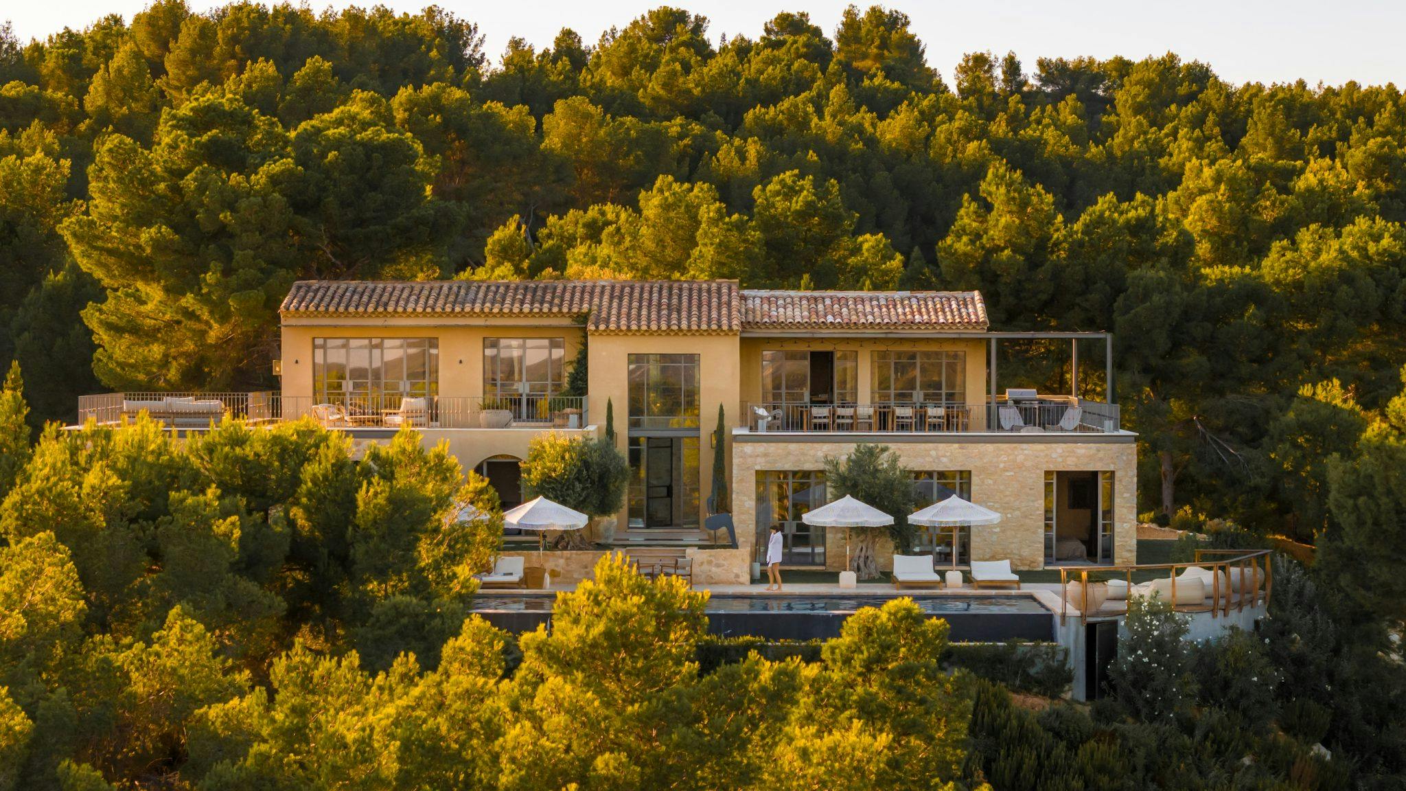 A large, luxurious villa with a terracotta roof sits amidst lush, green trees. The house features expansive glass doors and windows, multiple decks, outdoor seating areas with umbrellas, and a pool, all bathed in warm, golden sunlight.