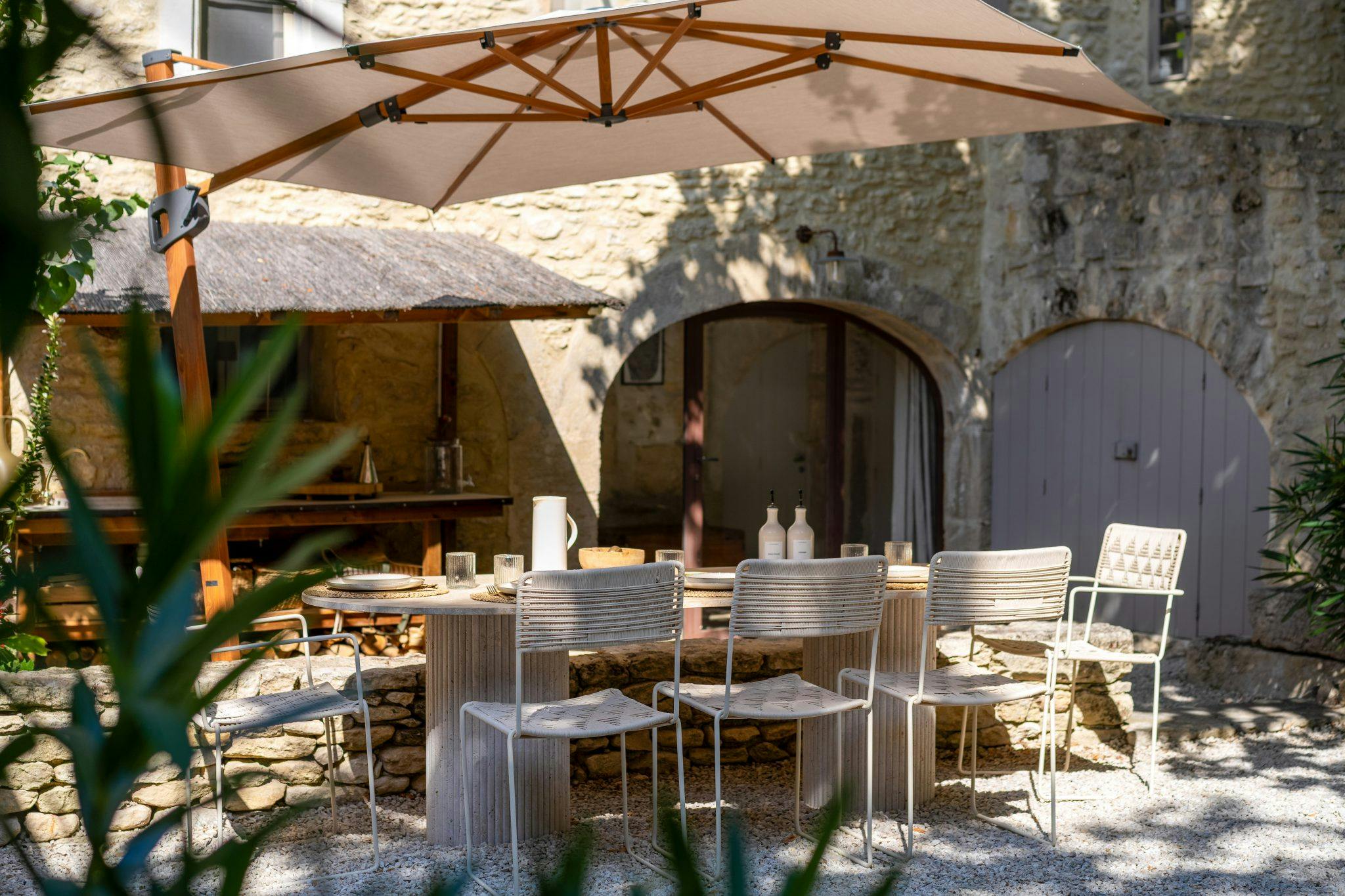 A quaint outdoor dining area is set with a rectangular wooden table, white chairs, and shaded by a large beige umbrella. The setting is rustic with stone walls, arched doorways, and greenery, creating a charming and inviting atmosphere.