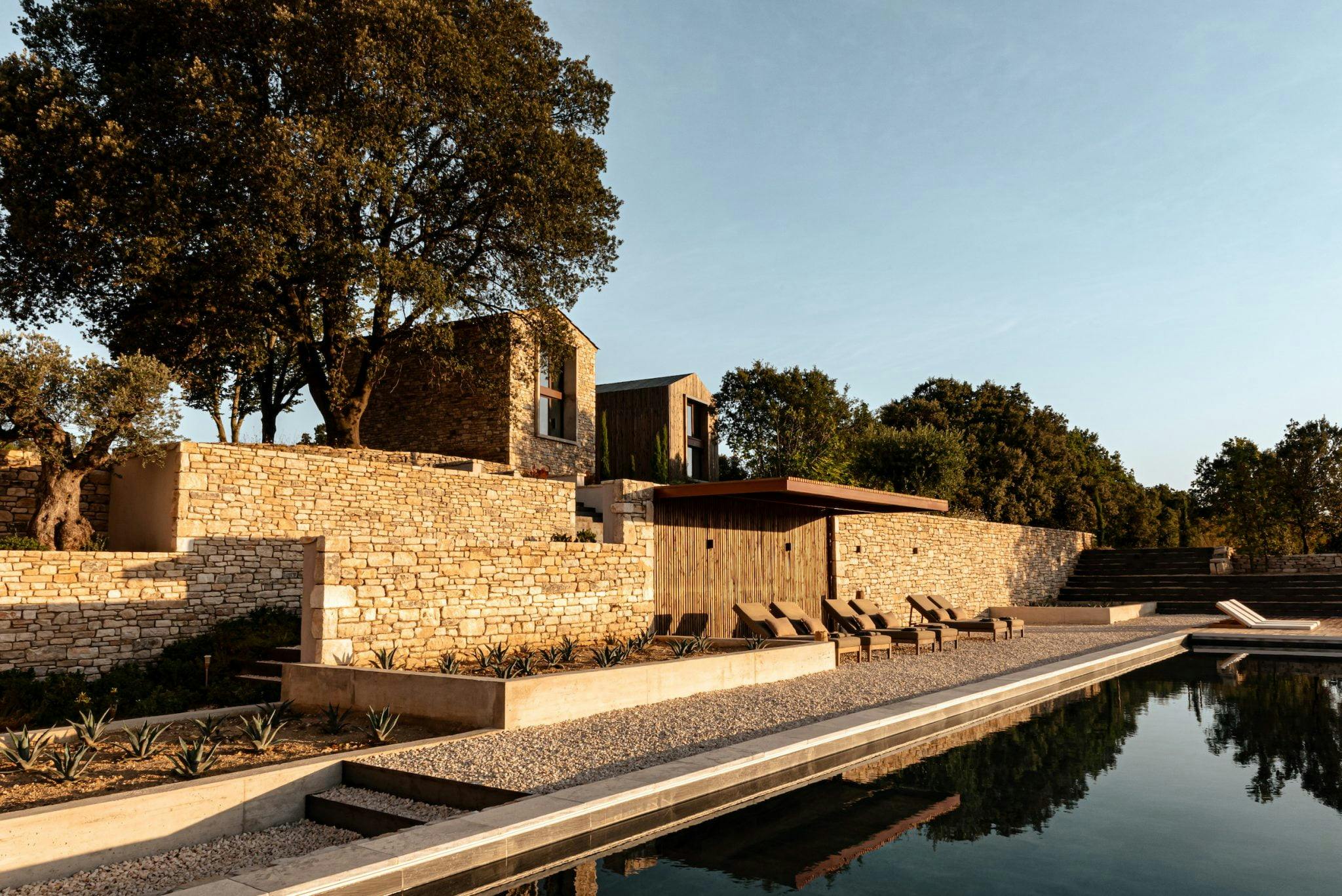 A modern villa with stone walls and large windows, situated on a hillside and surrounded by trees. In the foreground is a long rectangular swimming pool with sun loungers arranged on the pool deck. The scene is bathed in warm, late afternoon sunlight.