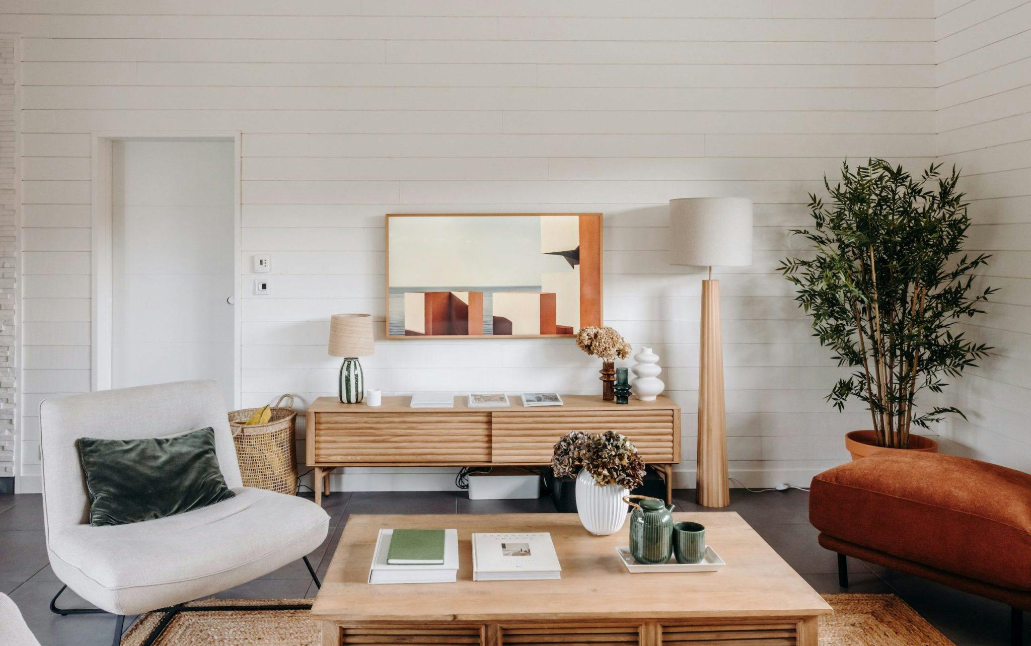 A cozy, modern living room with light wood furniture, including a low coffee table and a TV stand. There's a soft beige armchair, a rust-colored sofa, green plant, floor lamp, and decorative items such as books, vases, and a woven basket with a blanket.