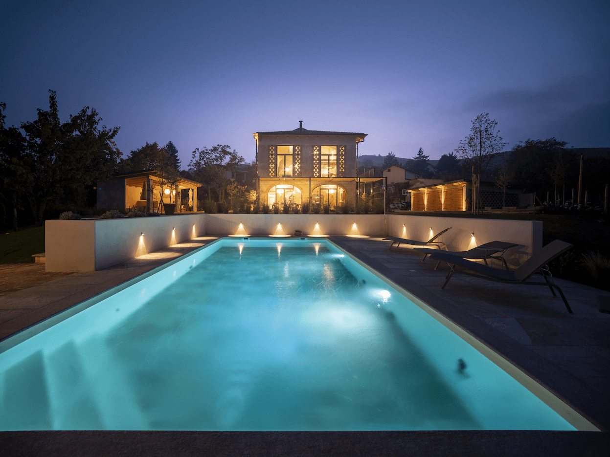 Une piscine luxueuse et éclairée la nuit est le point central au premier plan, menant à une maison moderne et bien éclairée à deux étages avec de grandes fenêtres et un patio. Autour de la piscine se trouvent des chaises longues et la toile de fond présente des arbres et un paysage serein.