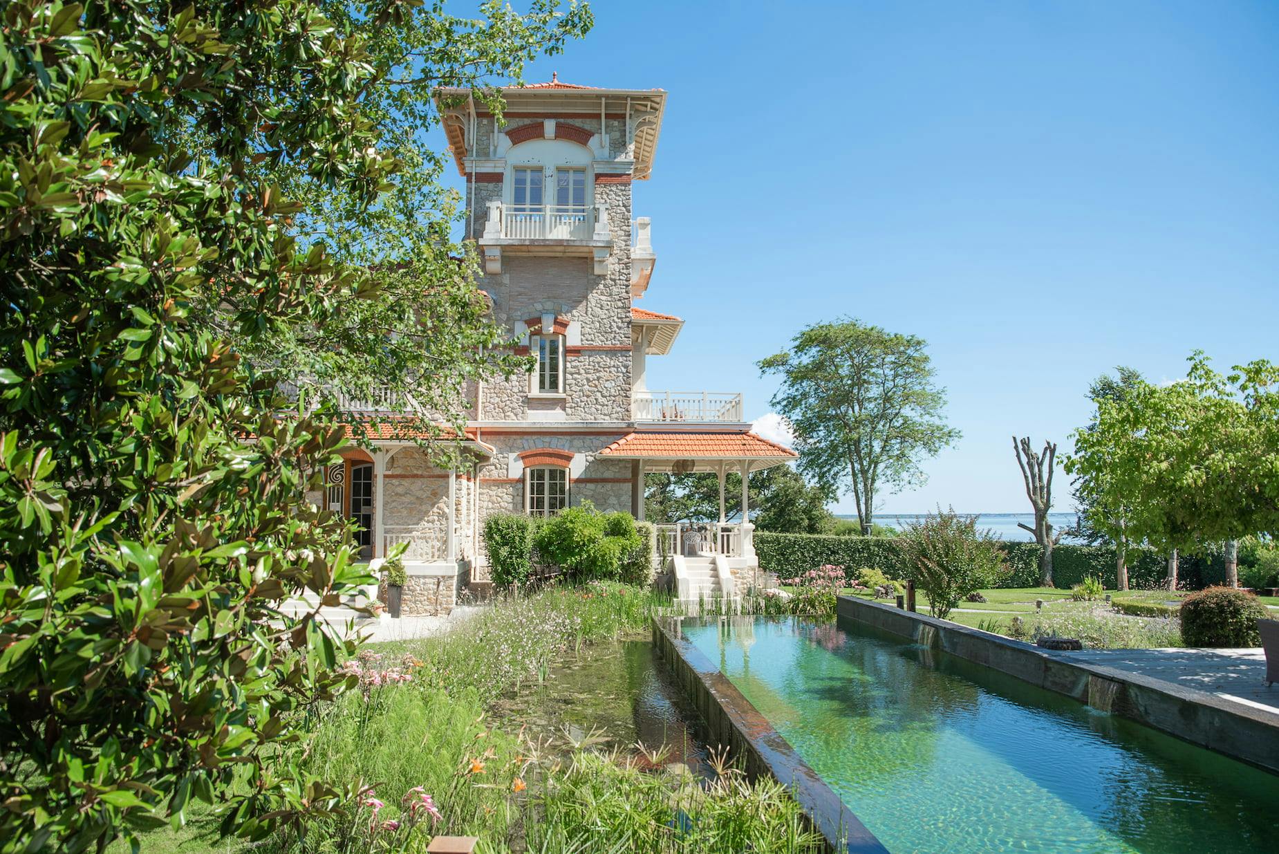 Une belle maison à plusieurs étages avec un extérieur en pierre et de grandes fenêtres est située dans un jardin verdoyant et luxuriant. Au premier plan, il y a un bassin réfléchissant long et étroit entouré de verdure. Le ciel est clair et bleu, renforçant l'atmosphère paisible.