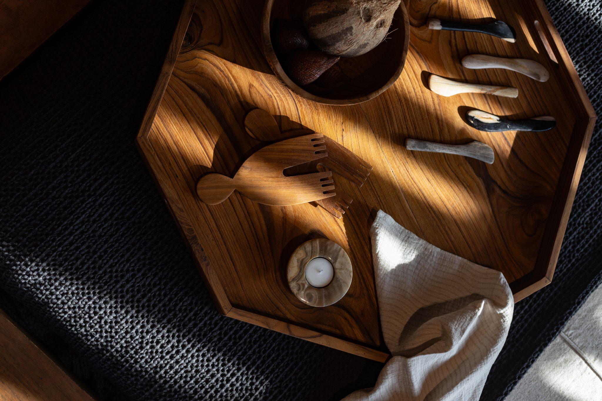 Un plateau en bois repose sur une surface texturée sombre, éclairée par la lumière naturelle. Sur le plateau se trouvent plusieurs objets dont trois petits couteaux aux manches légers, un objet en bois en forme de poisson, un bol et une petite bougie à côté d'un tissu blanc plié.