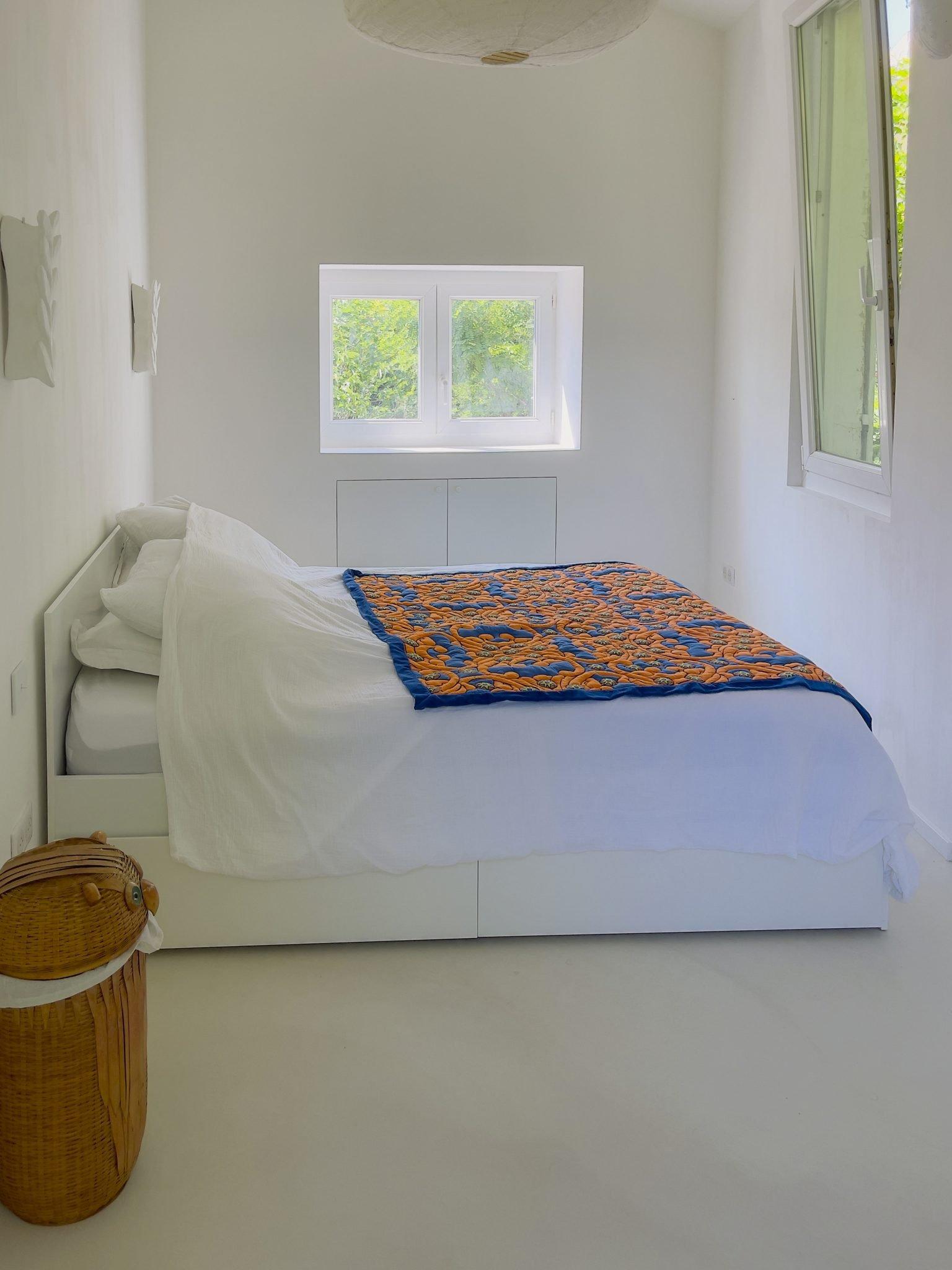 A minimalist bedroom features white walls and a white bed. The bed is adorned with a colorful blanket with blue and orange patterns. A small window and a large window allow natural light to fill the room. A woven laundry basket is placed next to the bed.