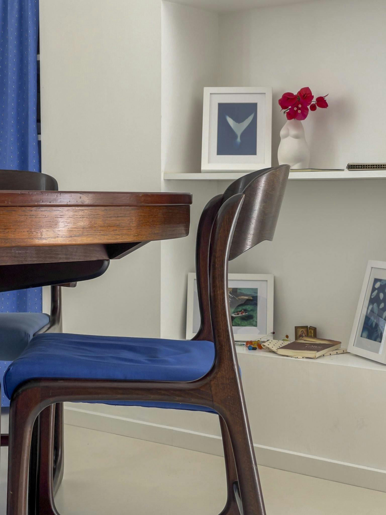 A dark-finished wooden dining table and a cushioned blue chair are in the foreground. In the background, white shelves display framed artwork, a vase with red flowers, and other decorative items. A blue curtain is partially visible on the left.