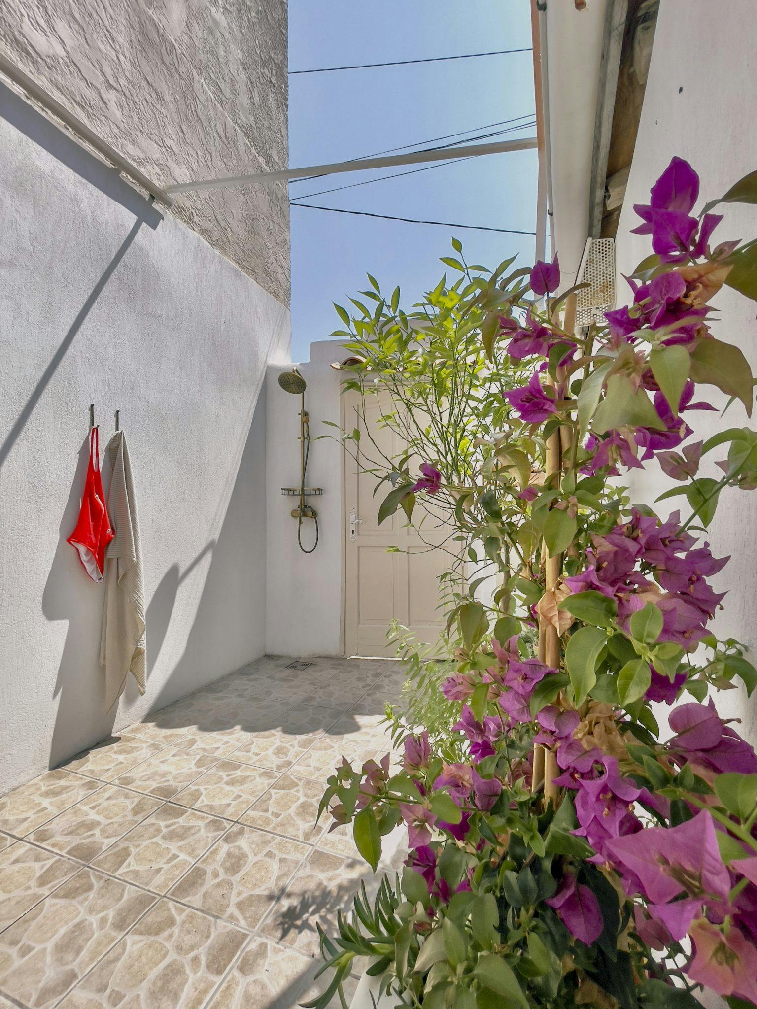 An outdoor shower area features white walls and a stone-tiled floor. Towels are hung on hooks to the left. A lush bougainvillea with vibrant purple flowers grows on the right. A showerhead and controls are mounted on the back wall under a clear blue sky.