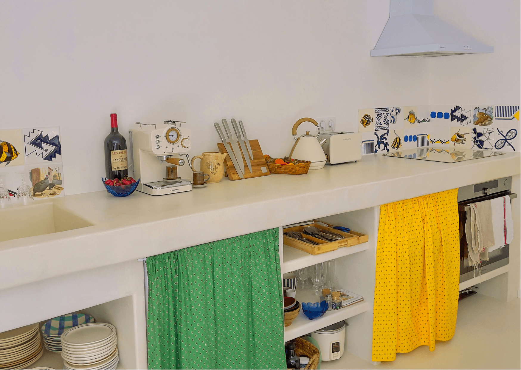 A well-lit kitchen includes a long white counter with various items, including a bottle of wine, a coffee machine, a blender, and a set of knives. Colorful tiles decorate the backsplash. The open shelves below store dishes and utensils, covered with green and yellow curtains.